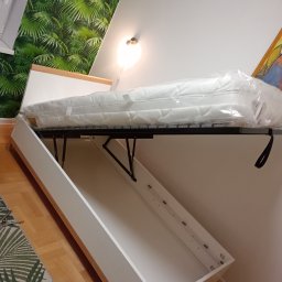 MULTI MONTI Janusz Zduńczyk - Rozkładane łóżko w szafie, materac jeszcze w folii, widoczne mechanizmy rozkładania, tapeta w tropikalne liście w tle, w rogu podłogi leżą szczypce.