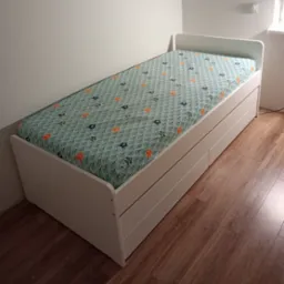 Białe łóżko jednoosobowe z szufladami, przykryte pościelą w morskie wzory, ustawione na drewnianej podłodze przy białej ścianie.