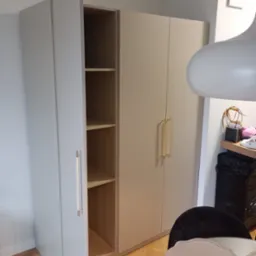 Jasnoszara szafa z otwartymi drzwiami ukazującymi puste półki, z długimi złotymi uchwytami, na tle parkietu i fragmentu pokoju z lampą.