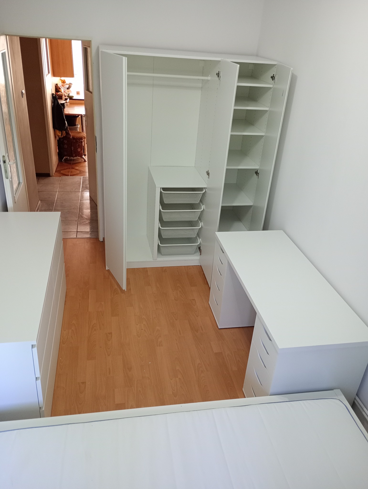 Biała szafa na wymiar z otwartymi drzwiami, półkami i koszami drucianymi, biurko z szufladami i komoda w pokoju z panelami podłogowymi.