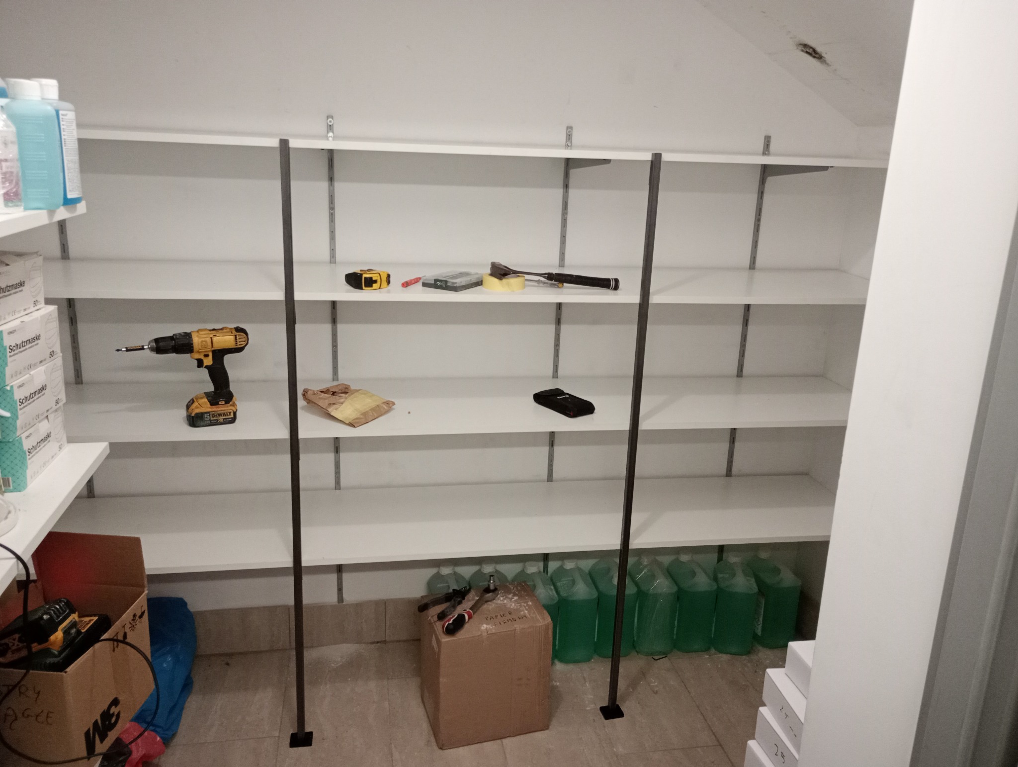 Białe półki zamontowane na ścianie, na których leżą narzędzia, akcesoria i materiały, w tym wiertarka akumulatorowa DeWalt, miara zwijana, poziomica, taśma klejąca, śrubokręt, opakowania maseczek...