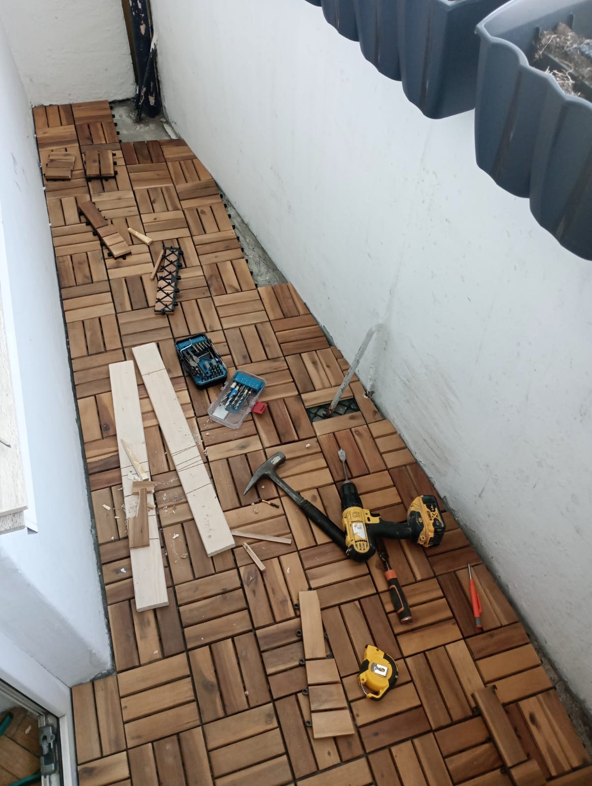 Montaż drewnianej podłogi z kwadratowych paneli na balkonie, widoczne narzędzia: wiertarka, młotek, miarka, śrubokręt i pudełka z wiertłami, częściowo ułożona podłoga.