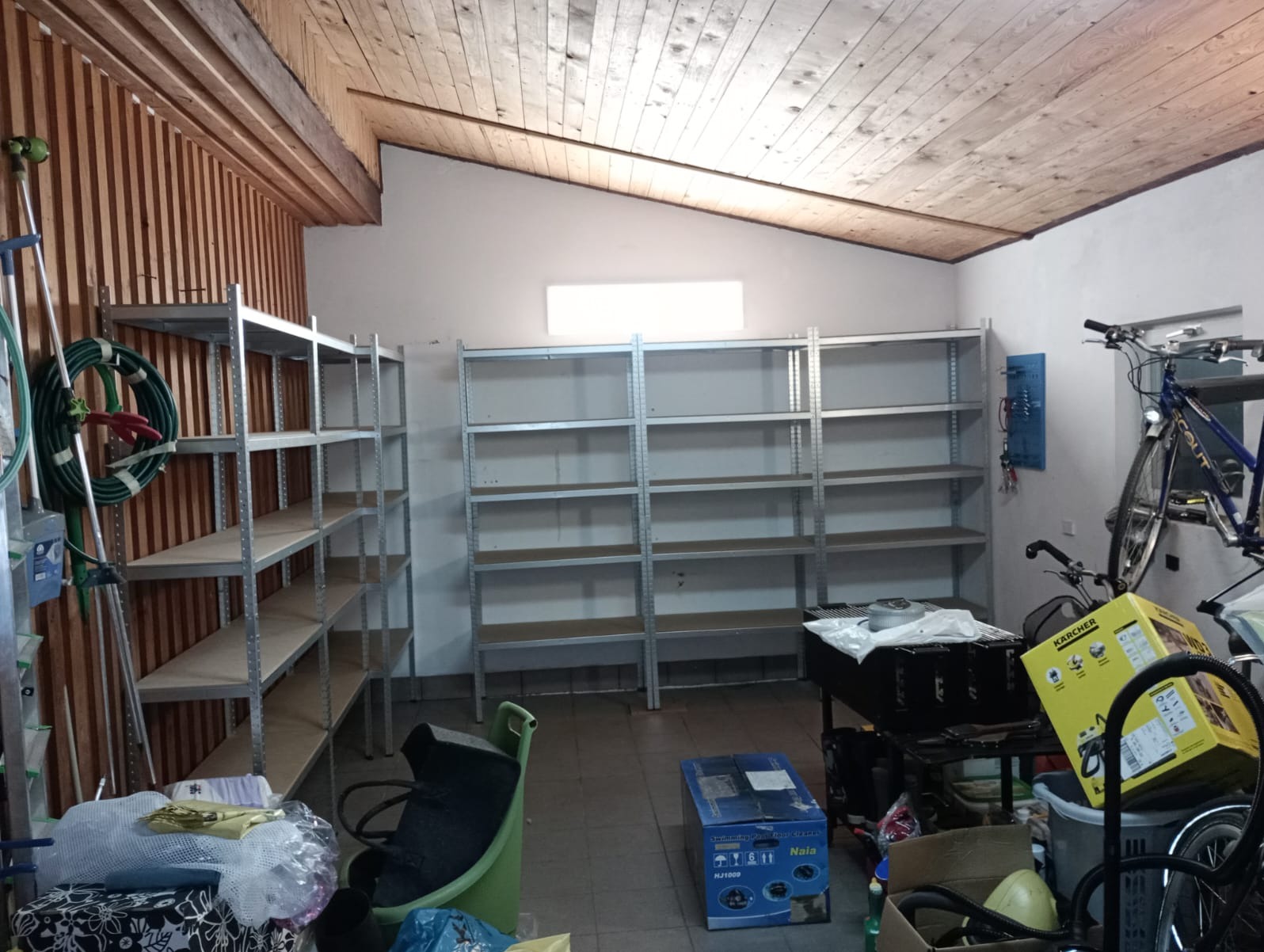 Metalowe regały magazynowe z półkami, częściowo puste, ustawione w garażu z drewnianym sufitem, widoczne narzędzia i sprzęty.