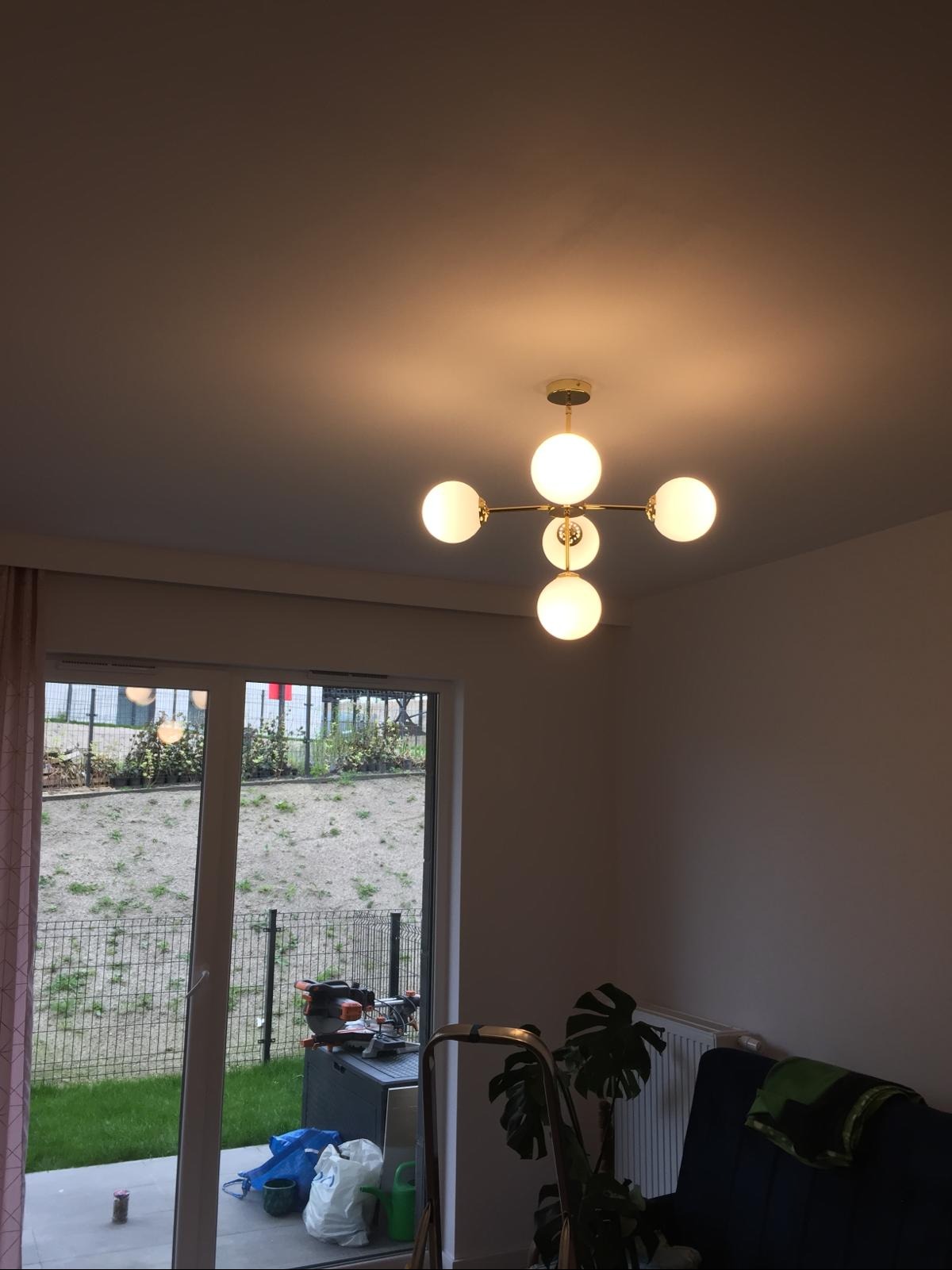 Nowoczesny żyrandol z pięcioma mlecznymi kulami zawieszony w pokoju, widok na zewnątrz przez okno, drabina oparta o ścianę, narzędzia widoczne na zewnątrz okna.