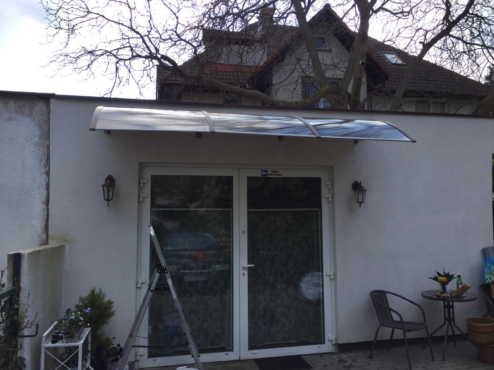 Zadaszenie nad dwuskrzydłowymi drzwiami wejściowymi, wsparte na białej ścianie budynku, z widoczną drabiną, lampami ściennymi i stolikiem z krzesłem na zewnątrz.