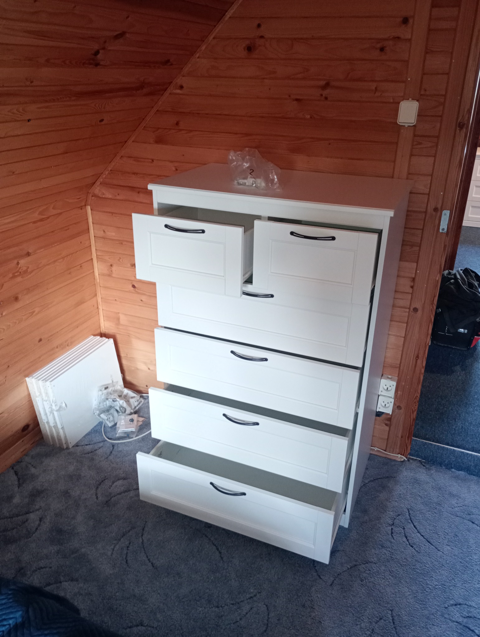 Biała komoda z otwartymi szufladami w pokoju wykończonym drewnianą boazerią, obok leżą elementy do montażu.