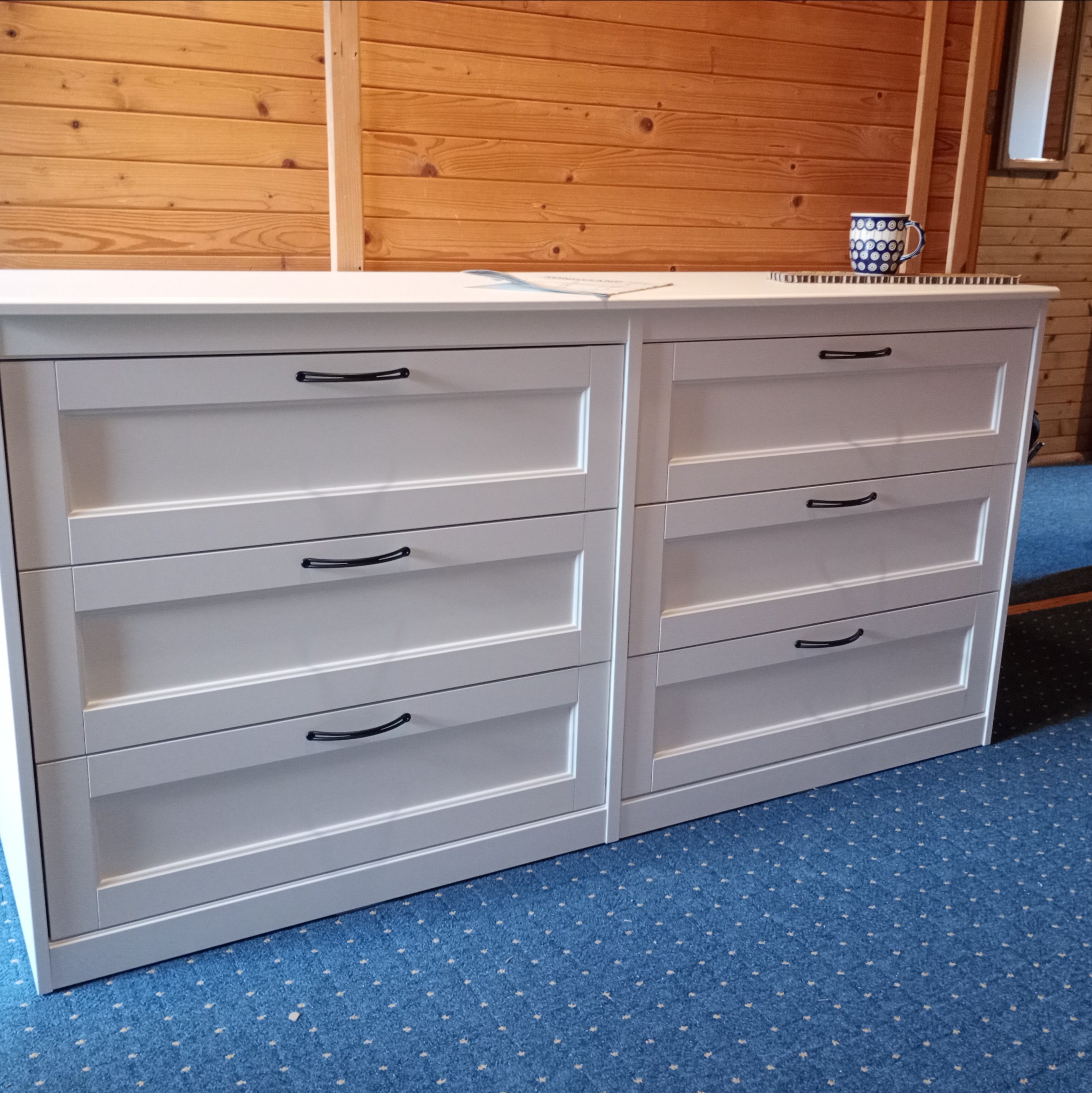 Biała komoda z sześcioma szufladami i czarnymi uchwytami, stojąca na niebieskiej wykładzinie, z drewnianą ścianą w tle, na której widać kubek i notatnik.