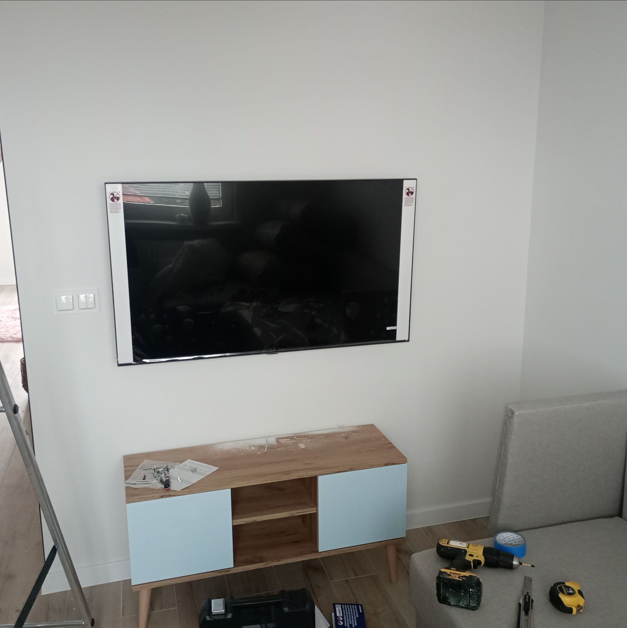 Telewizor zawieszony na białej ścianie nad komodą z błękitnymi drzwiczkami. Obok leży szara kanapa, a na niej wiertarka, taśma malarska i miarka. Na komodzie widoczne akcesoria montażowe, a po...