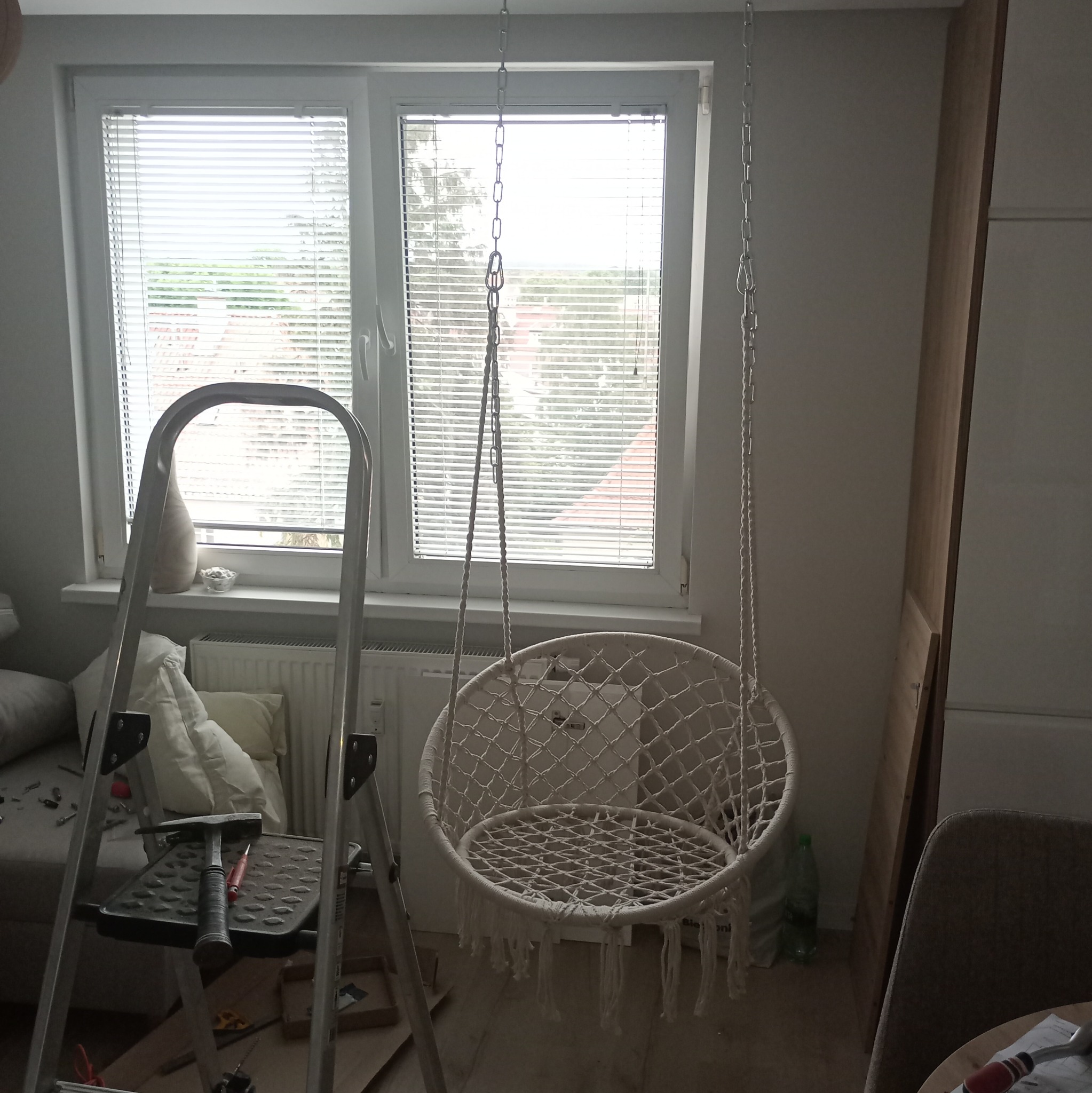 Białe, wiszące krzesło hamakowe z makramy zawieszone na łańcuchu przed oknem z żaluzjami, obok drabina z narzędziami, sugerujące prace montażowe w trakcie.