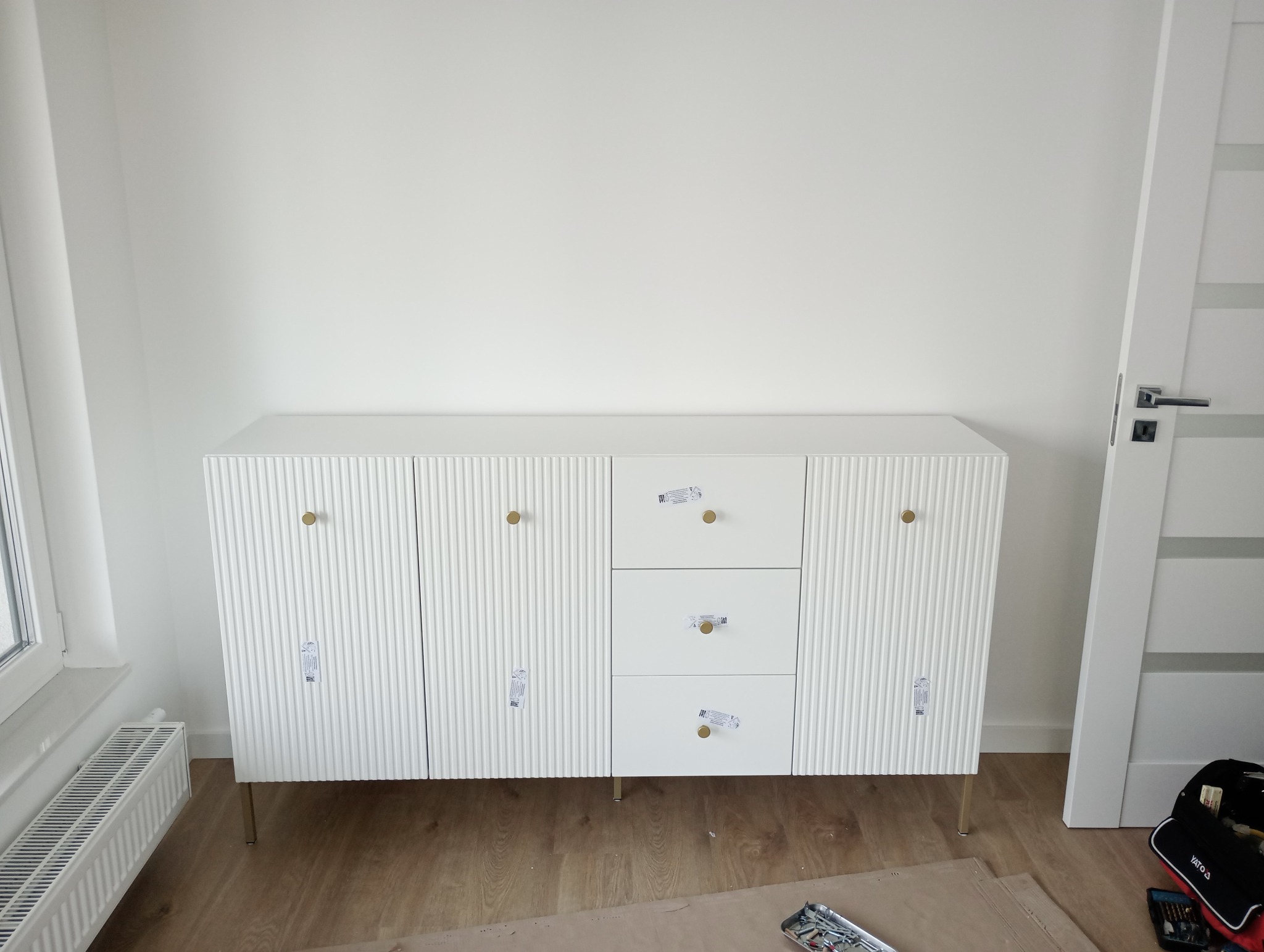 Biała komoda z trzema szufladami i dwojgiem drzwi z ryflowanym frontem, na złotych nóżkach, z naklejkami producenta, stoi na drewnianej podłodze przy białej ścianie, obok okna i białych drzwi.