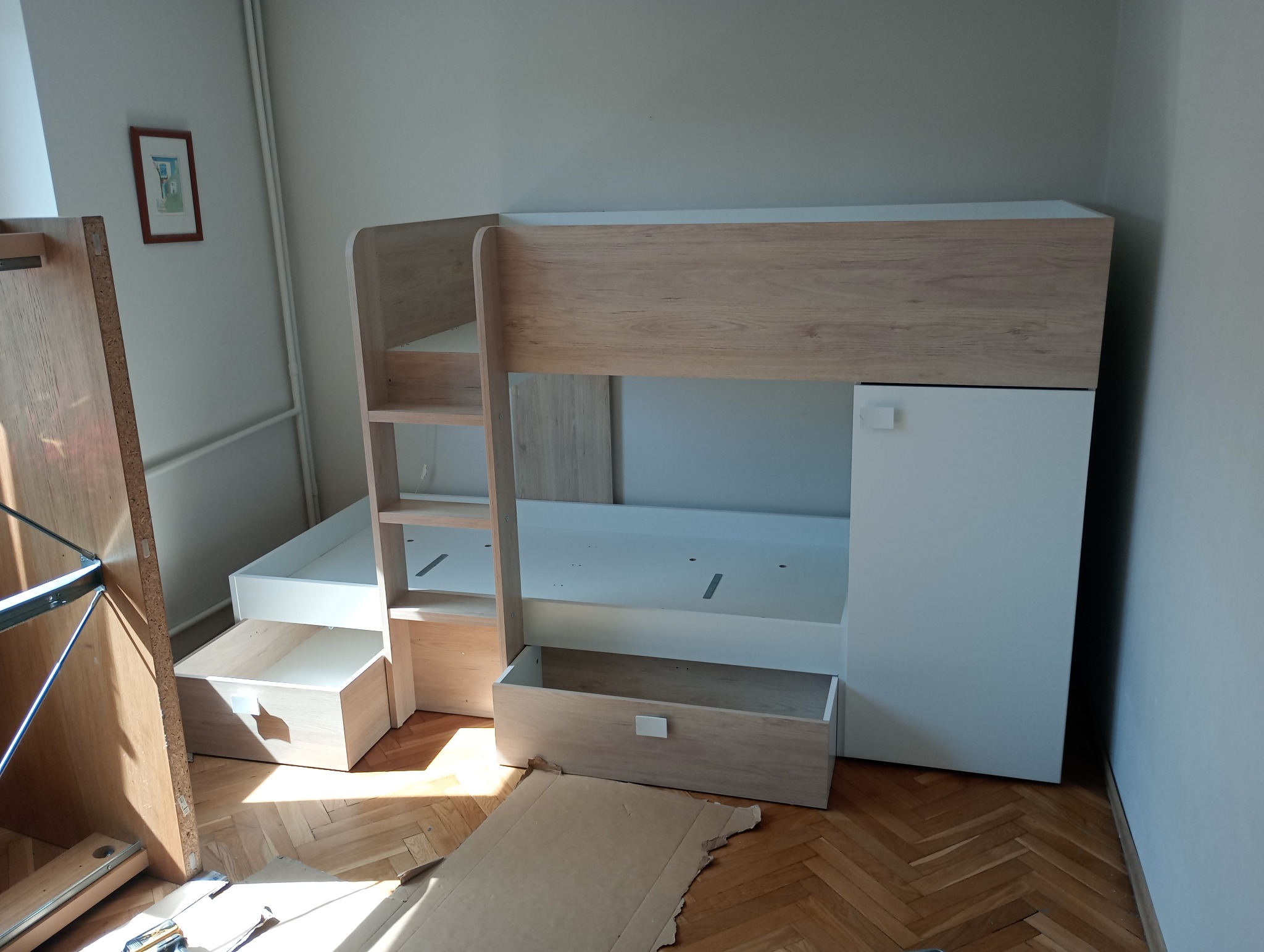Wysokie łóżko piętrowe z szufladami i szafką, częściowo zmontowane, z widocznymi elementami konstrukcyjnymi i kartonem na podłodze.