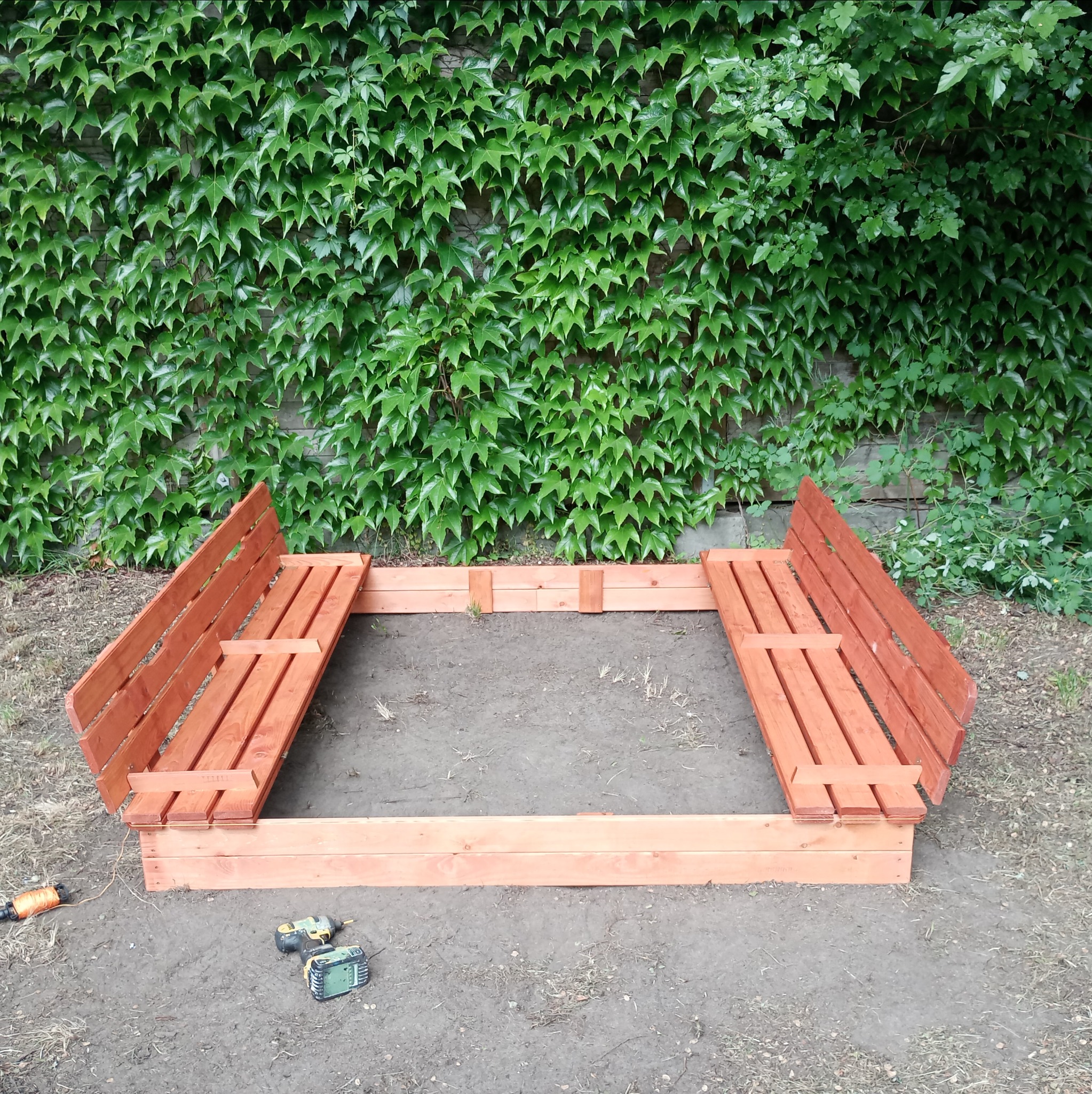 Drewniana piaskownica z wbudowanymi ławeczkami, częściowo wypełniona piaskiem, na tle ściany porośniętej bluszczem, z widocznymi narzędziami do montażu.