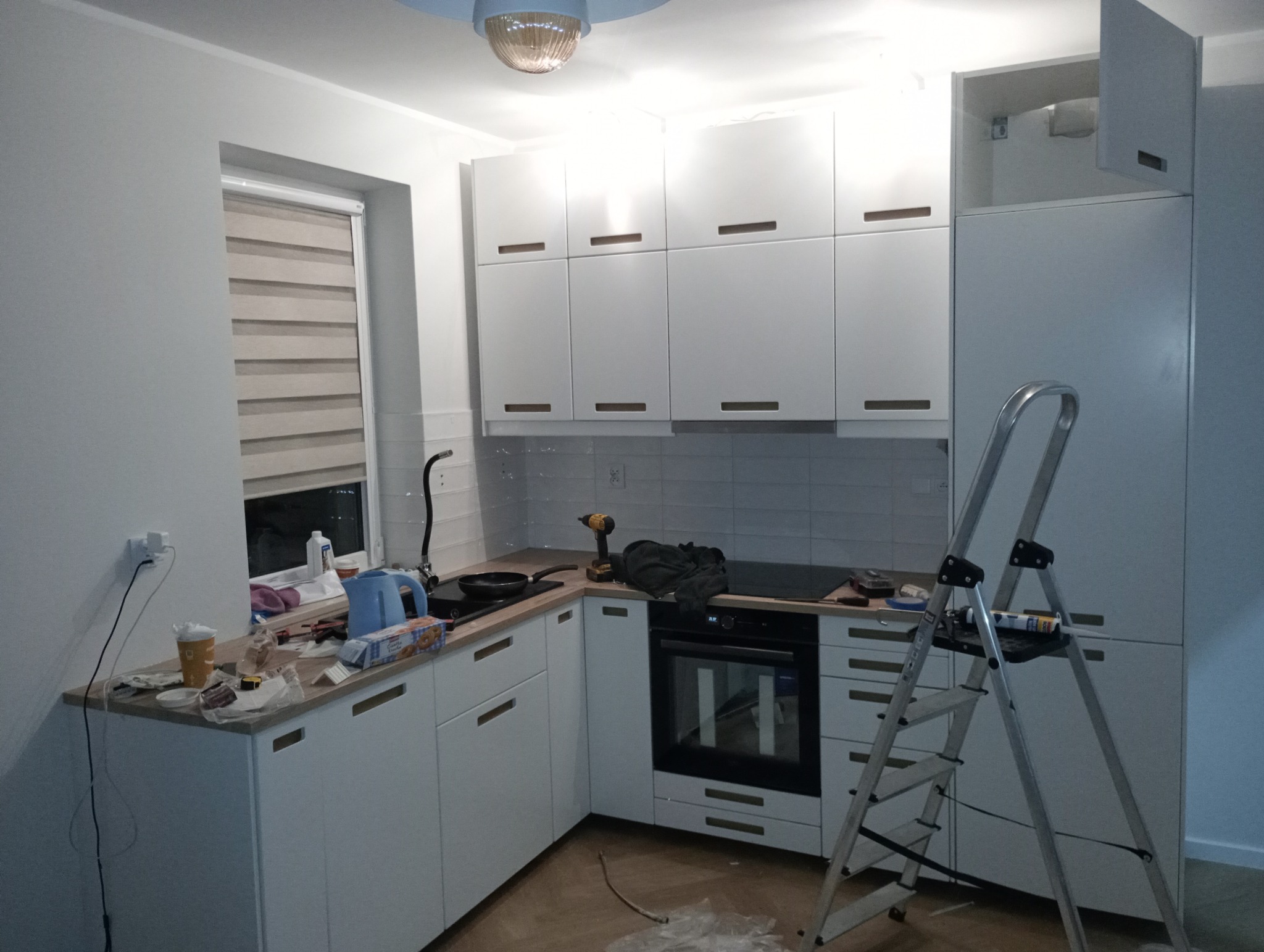 Wnętrze kuchni w trakcie montażu białych mebli kuchennych. Widoczna drabina, narzędzia i akcesoria montażowe na blacie. Część szafek zamontowana, pozostałe elementy czekają na instalację.