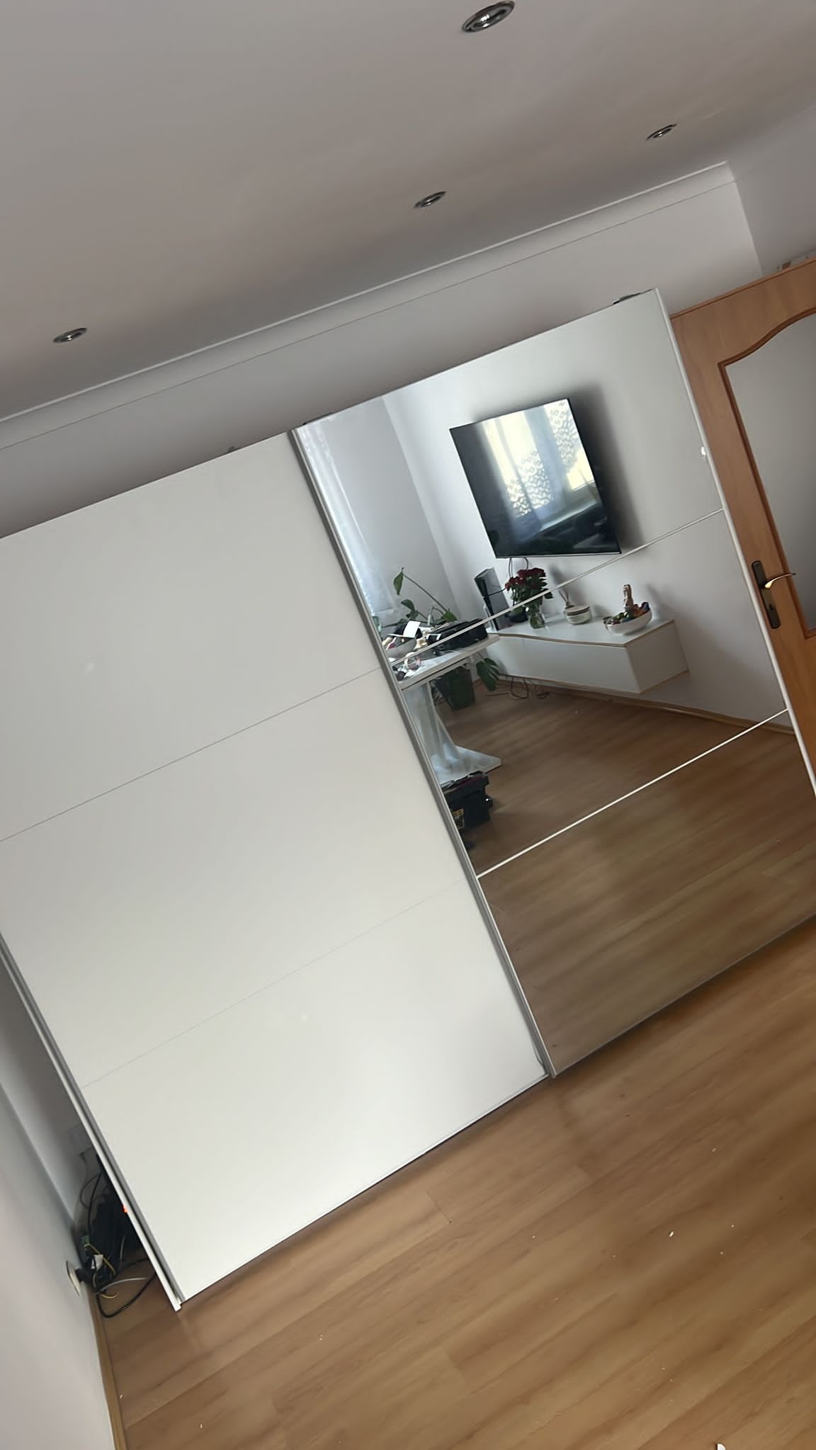 Nowoczesna szafa przesuwna z białymi frontami i lustrami, odbijająca wnętrze pokoju z telewizorem i drewnianą podłogą.