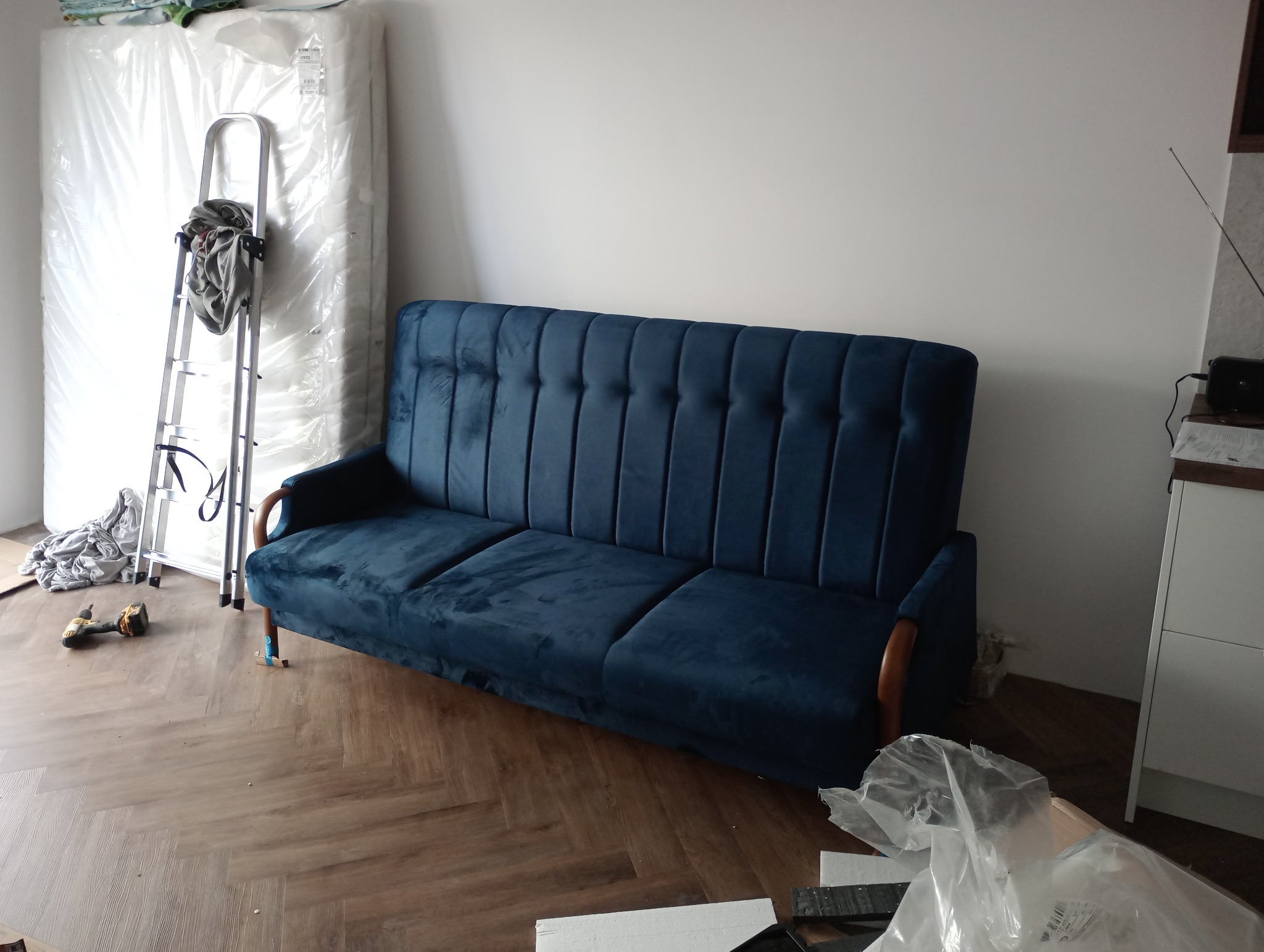 Niebieska, welurowa sofa z drewnianymi podłokietnikami, częściowo zmontowana, z wkrętarką i drabiną w tle, na parkiecie w jodełkę.