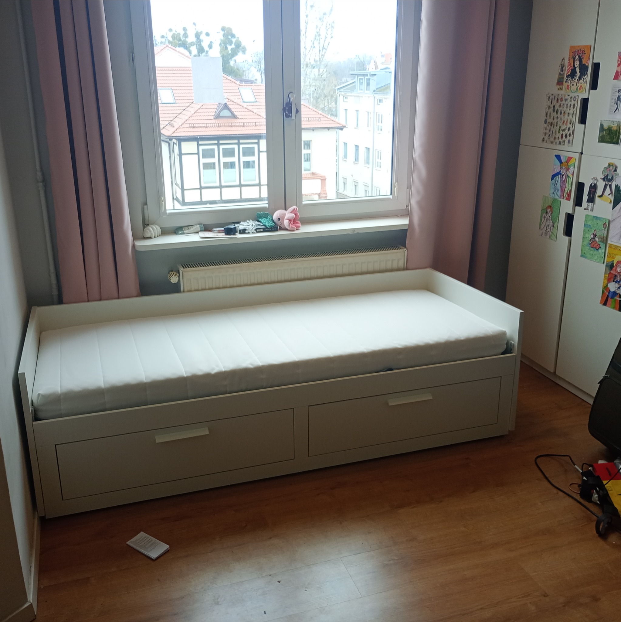 Białe łóżko z szufladami pod oknem, obok szafa z naklejkami, na podłodze panele i kartka.
