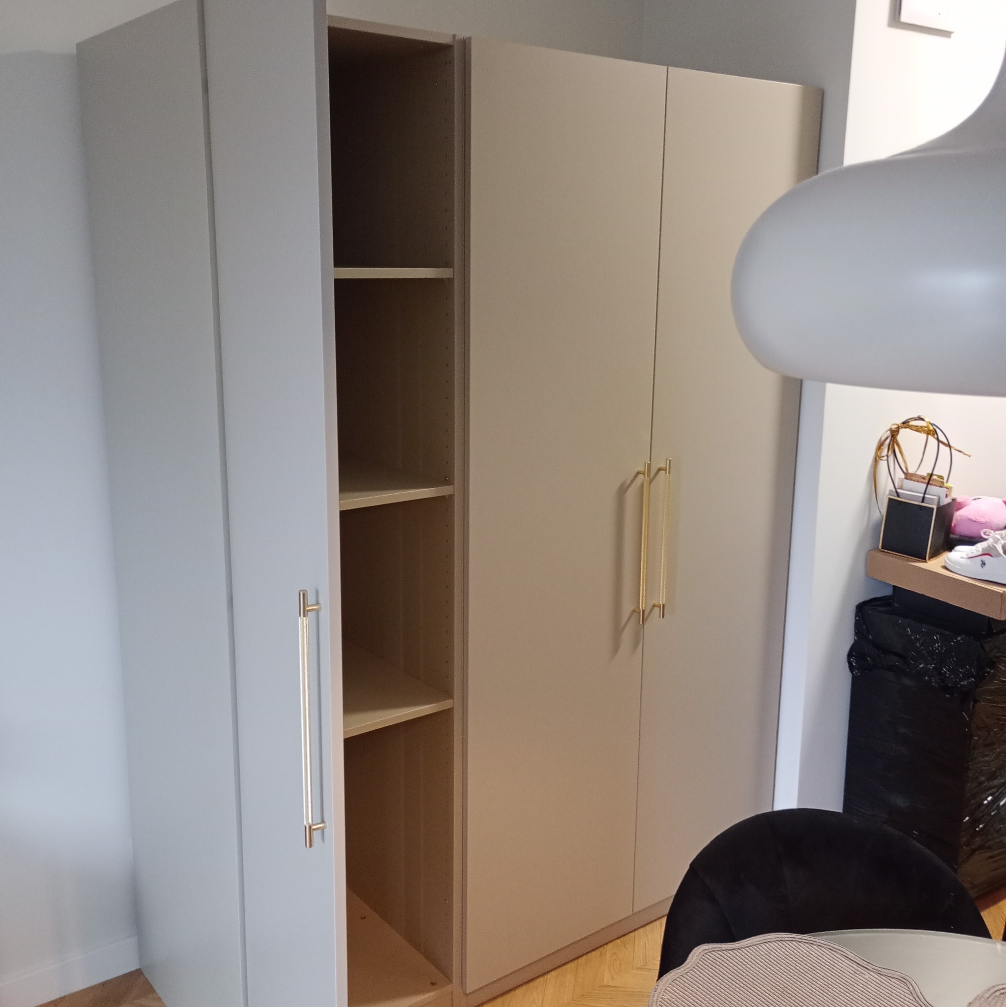 Jasnoszara szafa z otwartymi drzwiami ukazującymi puste półki, z długimi złotymi uchwytami, na tle parkietu i fragmentu pokoju z lampą.