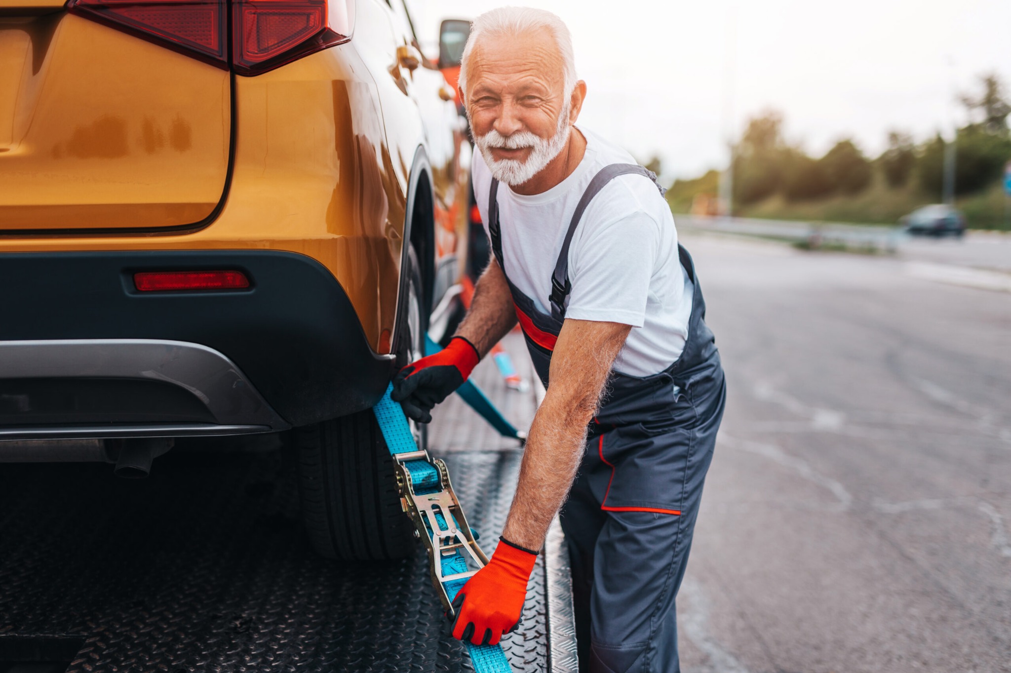 Uśmiechnięty mężczyzna w pomarańczowych rękawicach zabezpiecza pomarańczowy samochód pasami na platformie lawety, widoczny fragment drogi w tle.