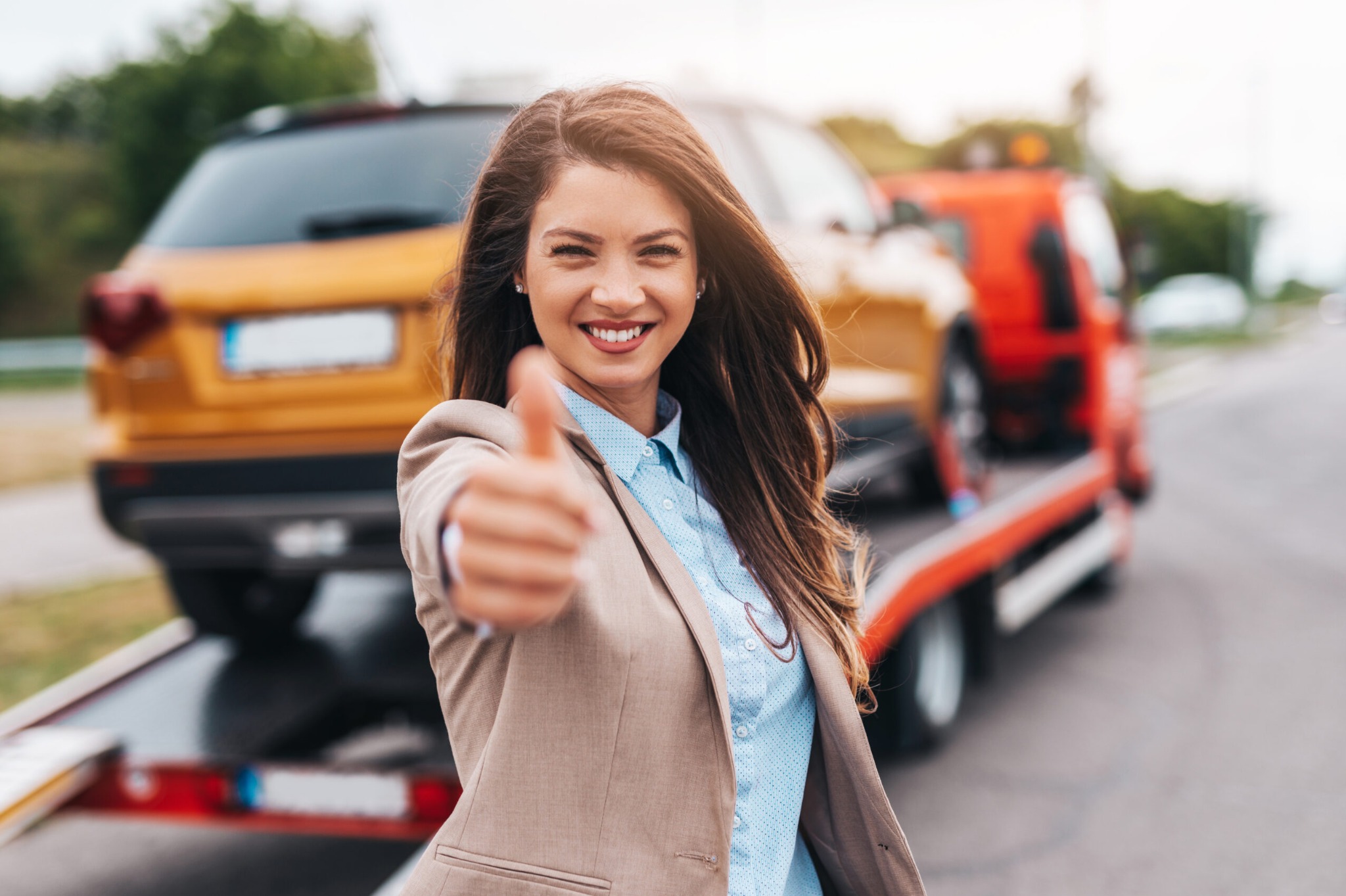 Uśmiechnięta kobieta w beżowej marynarce pokazuje kciuk w górę na tle lawety z przewożonym żółtym SUV-em.