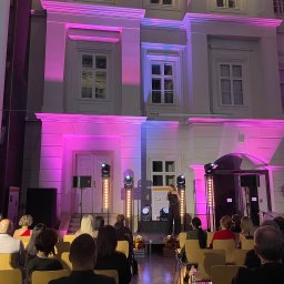 GRUPA L8 SPÓŁKA Z OGRANICZONĄ ODPOWIEDZIALNOŚCIĄ - Prezentacja na scenie przed budynkiem oświetlonym na różowo, widownia siedzi na krzesłach.