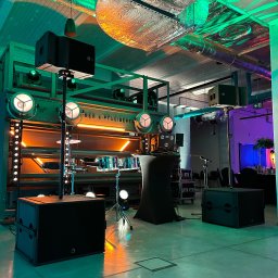 GRUPA L8 SPÓŁKA Z OGRANICZONĄ ODPOWIEDZIALNOŚCIĄ - Scena z bębnami, oświetleniem punktowym i nagłośnieniem w industrialnym wnętrzu, z widocznymi elementami konstrukcyjnymi budynku i instalacjami na suficie, oświetlonymi kolorowymi światłami.