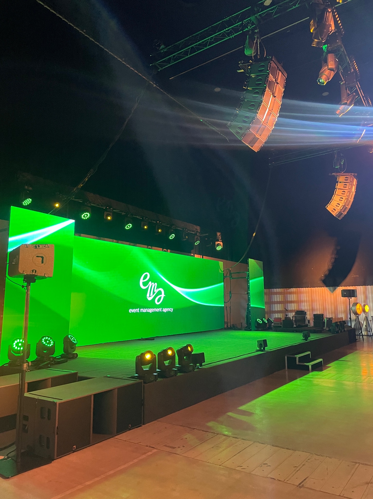 Scena z zielonym ekranem LED wyświetlającym logo 'ema event management agency', oświetlona reflektorami, z wiszącym systemem nagłośnieniowym line array oraz dodatkowym oświetleniem scenicznym.