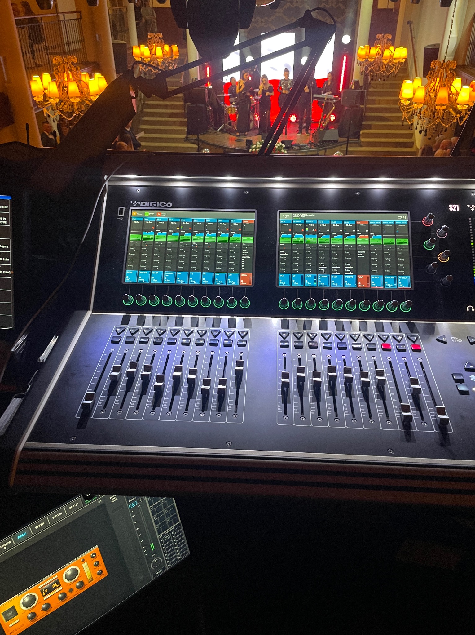 Konsoleta mikserska DiGiCo z widokiem na scenę z zespołem muzycznym i dekoracyjnymi żyrandolami w tle.