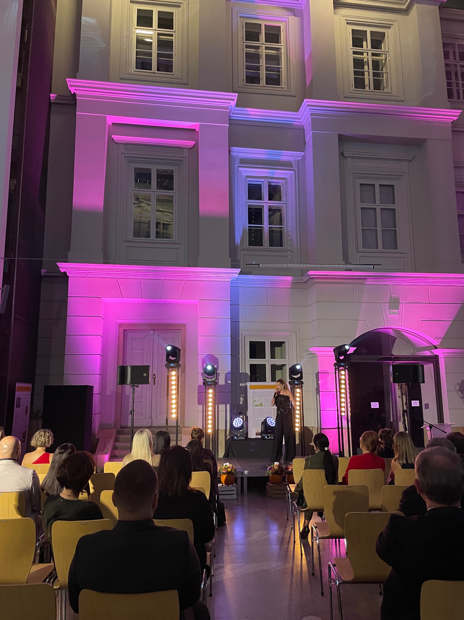 Prezentacja na scenie przed budynkiem oświetlonym na różowo, widownia siedzi na krzesłach.