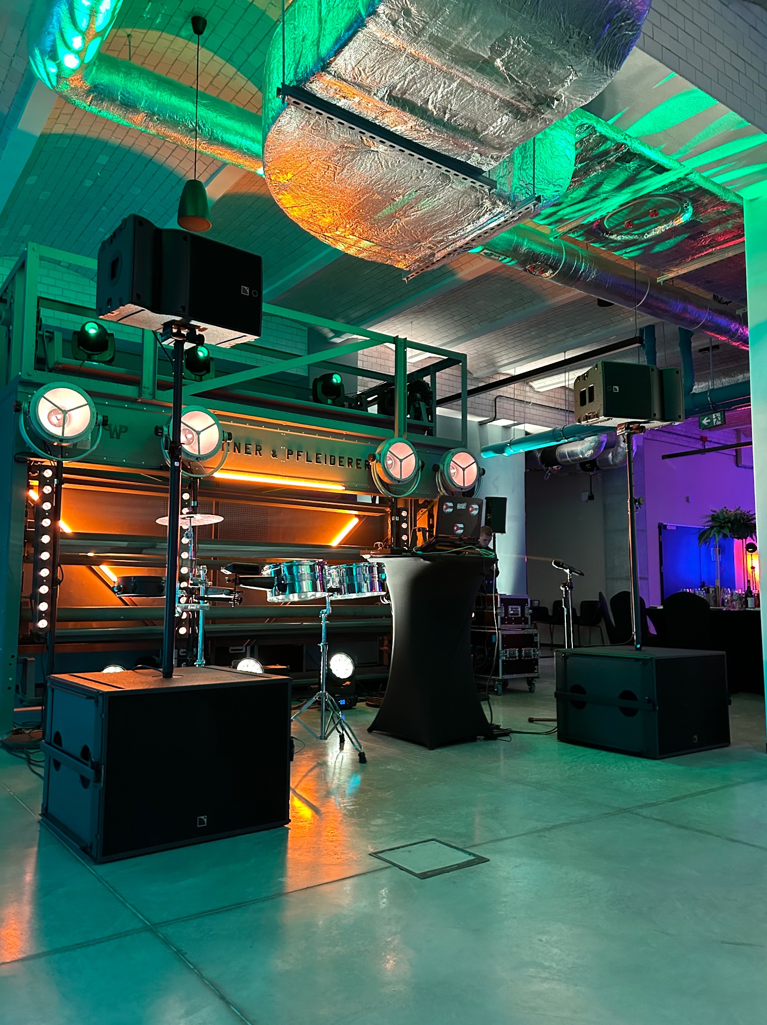 Scena z bębnami, oświetleniem punktowym i nagłośnieniem w industrialnym wnętrzu, z widocznymi elementami konstrukcyjnymi budynku i instalacjami na suficie, oświetlonymi kolorowymi światłami.