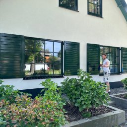 Usługi Budowlane Artur Jedrzejewski - Mężczyzna myje okna w domu z zielonymi okiennicami, widoczne rośliny w ogródku przed domem.