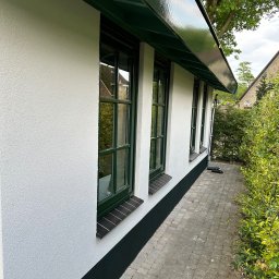 Usługi Budowlane Artur Jedrzejewski - Ściana budynku z oknami w zielonych ramach, po świeżym malowaniu na biało z zielonymi akcentami na podmurówce i podbitce dachu, widok z zewnątrz na tle kostki brukowej i zieleni.