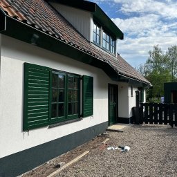 Usługi Budowlane Artur Jedrzejewski - Elewacja domu z zielonymi okiennicami i pomalowaną na ciemno dolną częścią ściany, widoczne narzędzia i materiały malarskie na żwirowym podłożu.