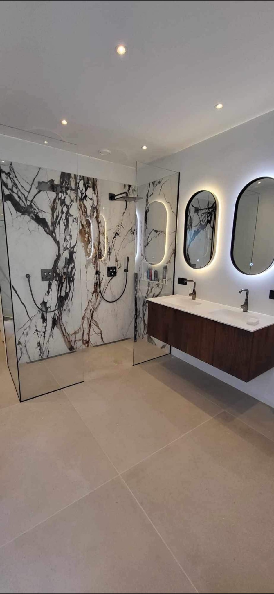 Nowoczesna łazienka z kabiną prysznicową typu walk-in wykończoną płytami imitującymi marmur z czarnymi żyłami, podwieszaną szafką z umywalką nablatową i owalnymi lustrami z podświetleniem LED.