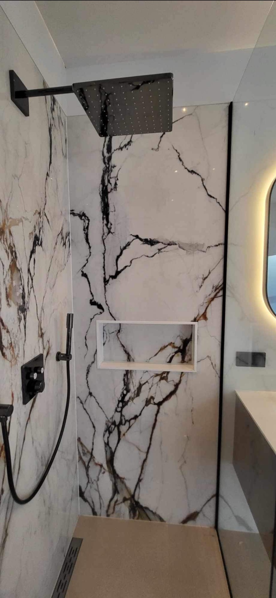 Nowoczesna kabina prysznicowa z marmurowymi płytkami, czarną armaturą i wnęką na kosmetyki.