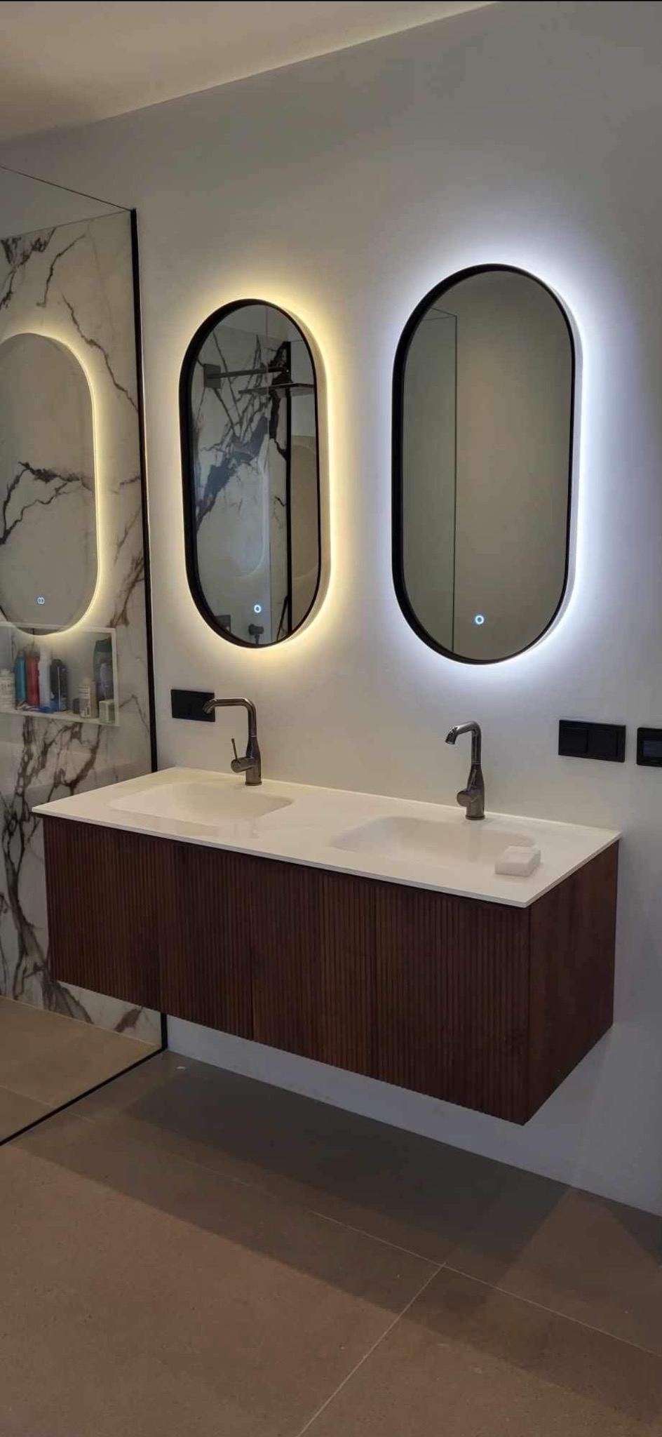 Nowoczesna łazienka z podwójną umywalką na drewnianej szafce, owalne lustra z podświetleniem LED i marmurowa ściana w tle.