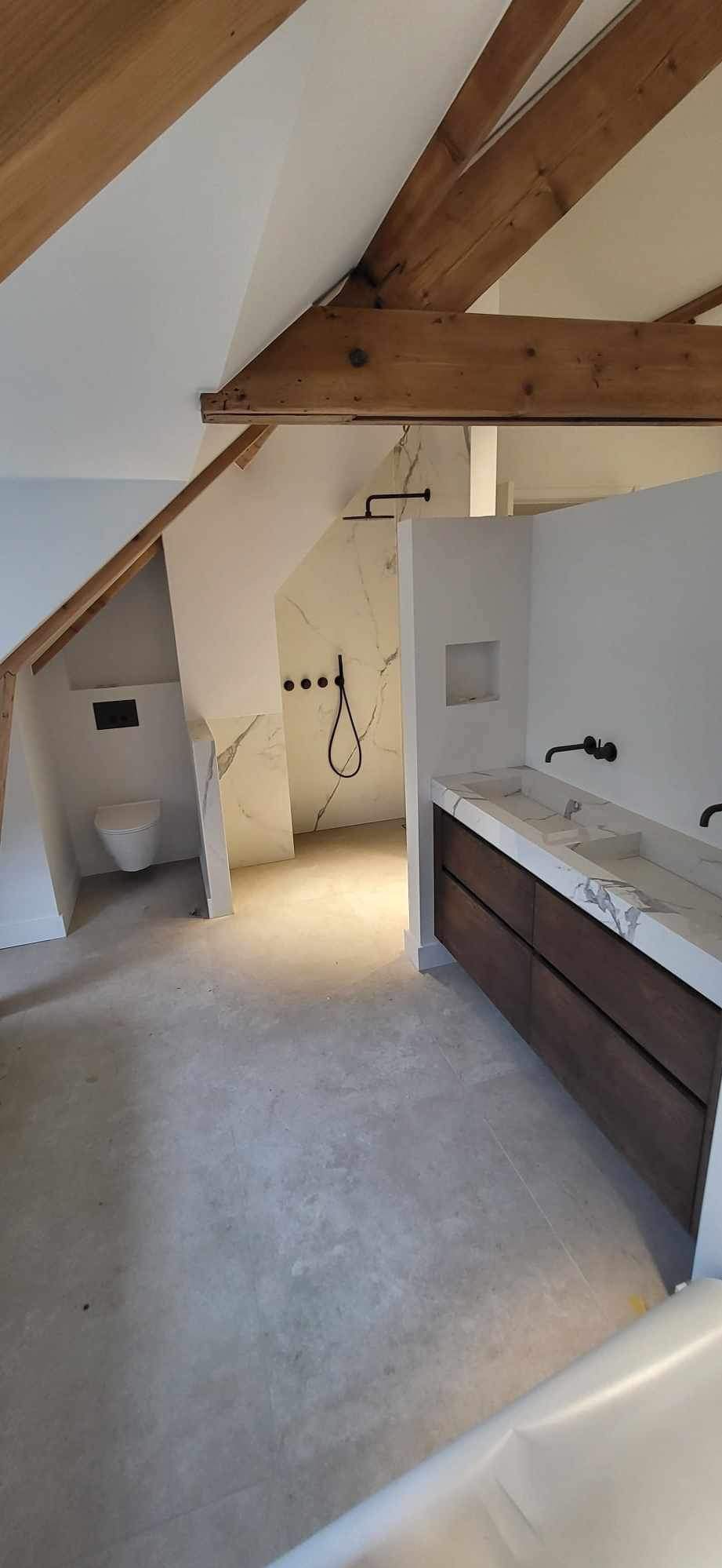 Nowoczesna łazienka na poddaszu z drewnianymi belkami stropowymi, marmurowymi płytami pod prysznicem i umywalką oraz minimalistyczną armaturą w kolorze czarnym.