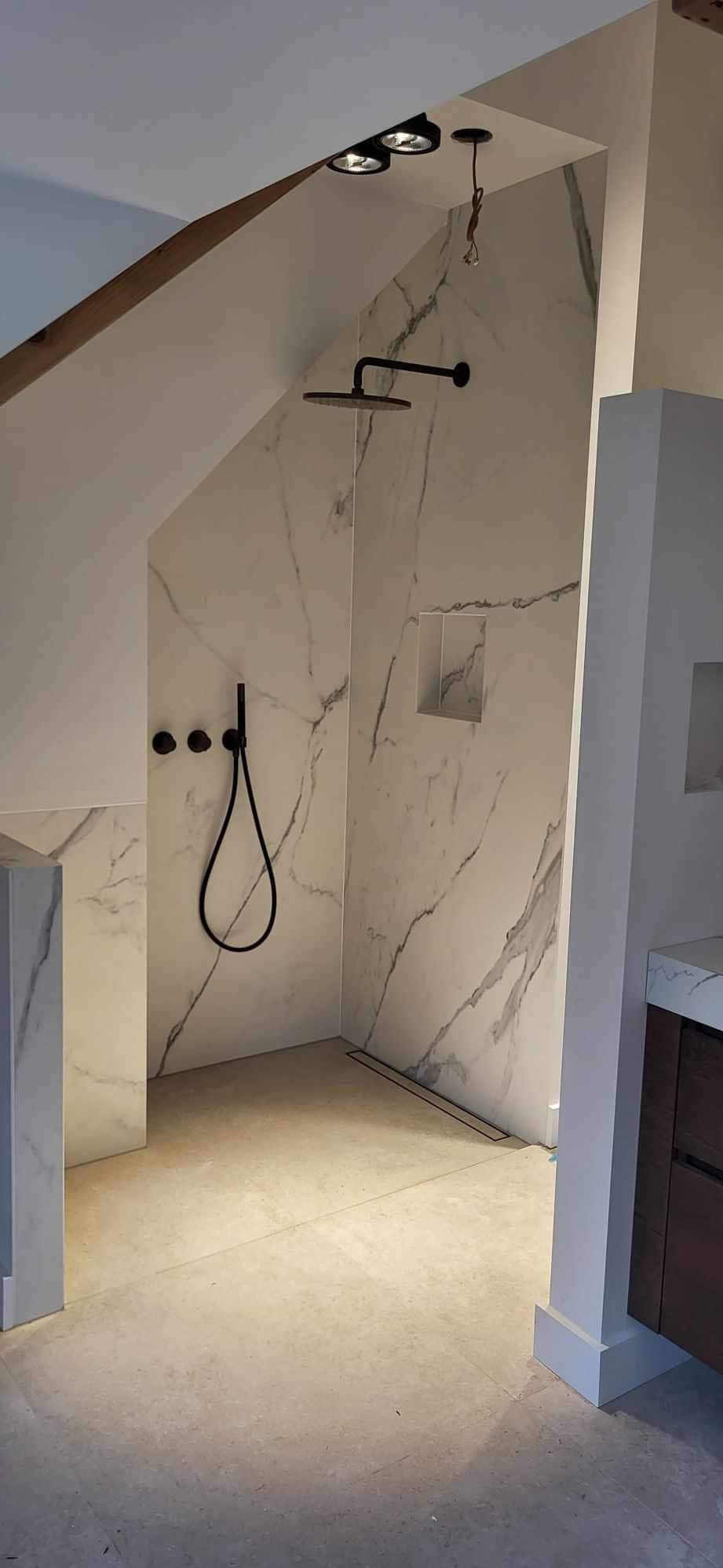 Nowoczesna kabina prysznicowa z marmurowymi płytami, czarna armatura i punktowe oświetlenie w skośnej łazience.