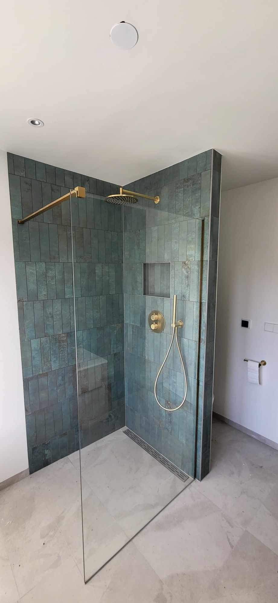 Nowoczesna kabina prysznicowa walk-in z zielono-niebieskimi płytkami wertykalnymi i złotą armaturą, widoczny odpływ liniowy w podłodze z jasnego gresu.