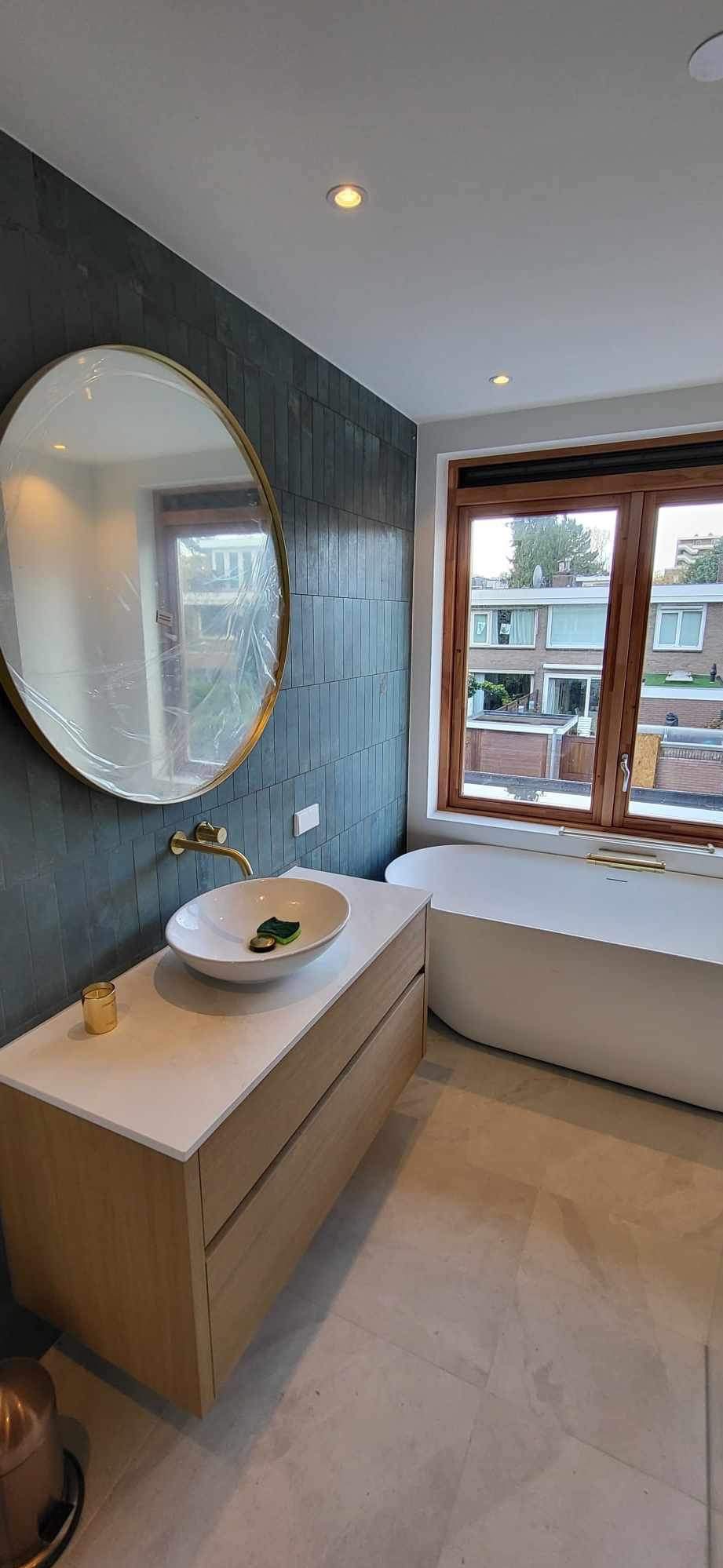 Wykończona łazienka z ciemnymi, pionowo ułożonymi płytkami, owalnym lustrem w złotej ramie, białą umywalką nablatową, drewnianą szafką pod umywalką i wolnostojącą wanną przy oknie z drewnianą ramą.