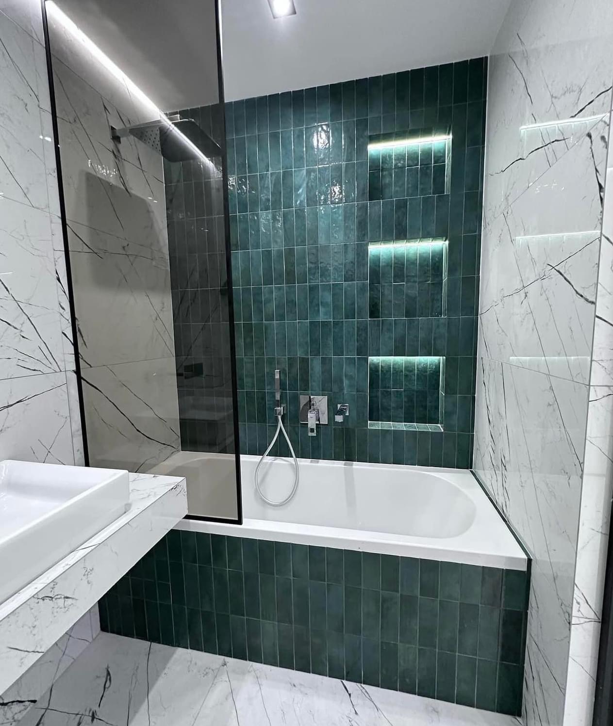 Nowoczesna łazienka z zielonymi płytkami ułożonymi pionowo, podświetlane wnęki w ścianie, kabina prysznicowa z czarną ramą i marmurowy blat umywalkowy.