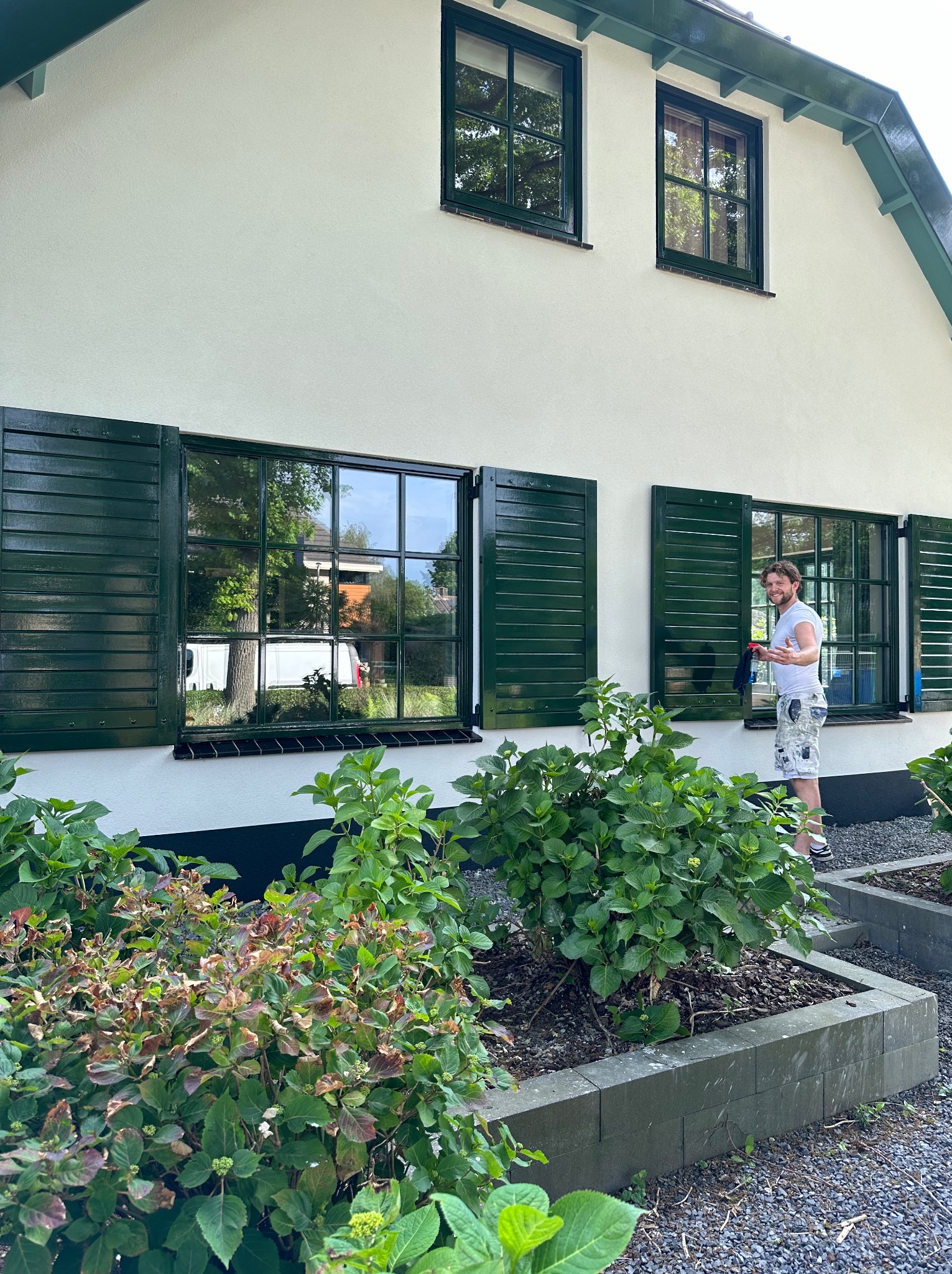 Mężczyzna myje okna w domu z zielonymi okiennicami, widoczne rośliny w ogródku przed domem.