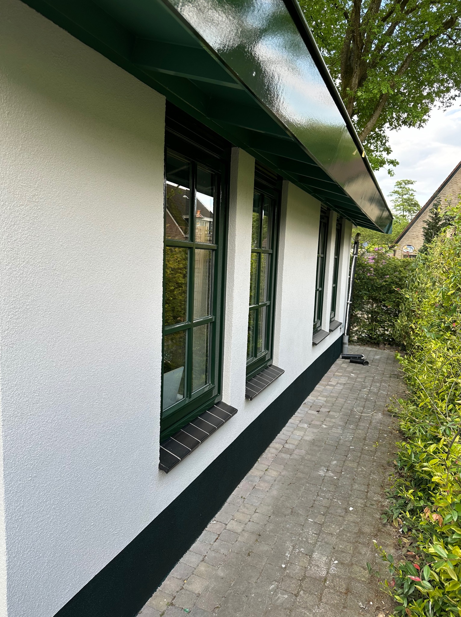 Ściana budynku z oknami w zielonych ramach, po świeżym malowaniu na biało z zielonymi akcentami na podmurówce i podbitce dachu, widok z zewnątrz na tle kostki brukowej i zieleni.