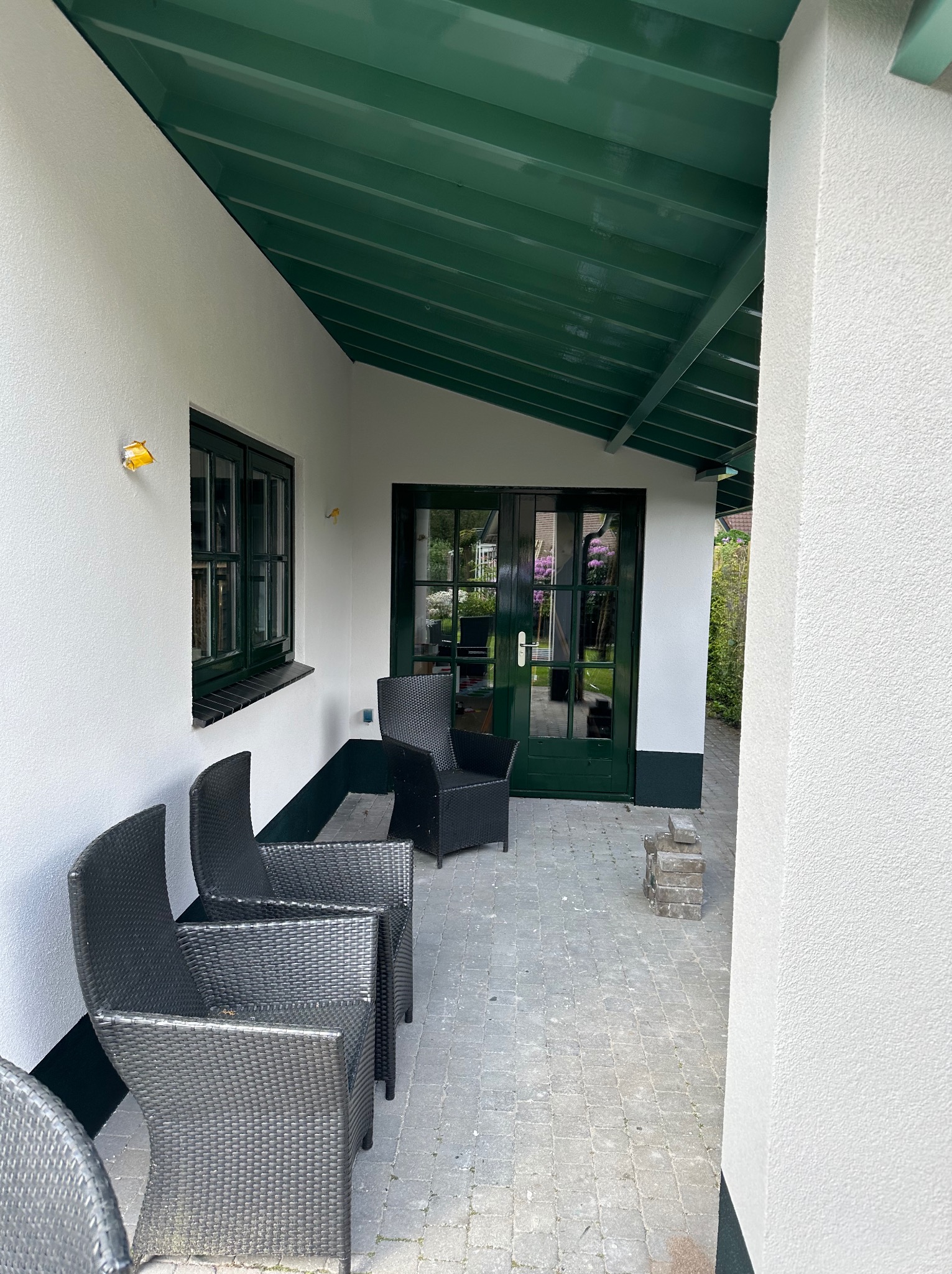 Kryty taras z białymi ścianami, zielonym sufitem i ramami okiennymi, wyposażony w cztery wiklinowe krzesła i przeszklone drzwi.