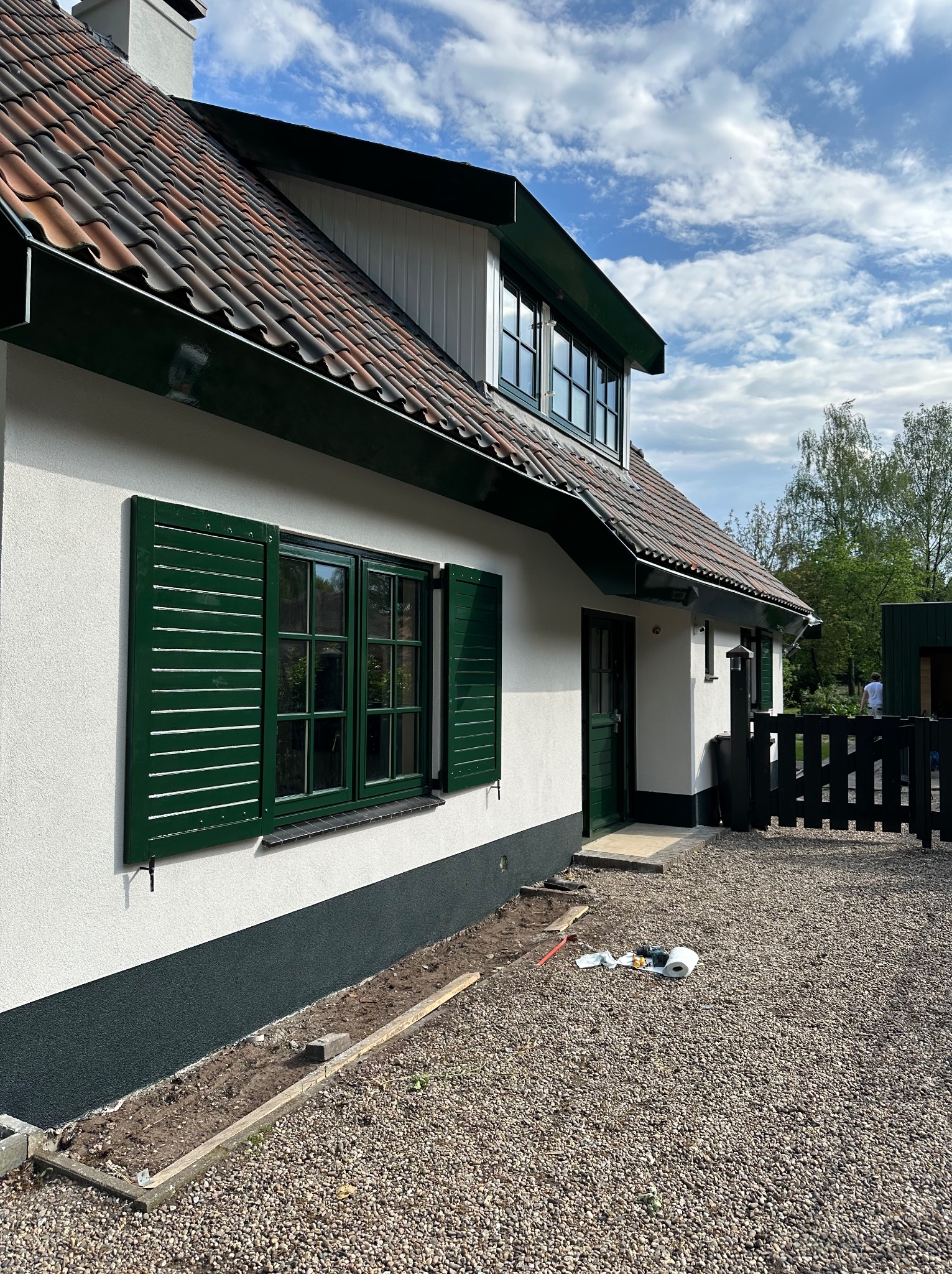 Elewacja domu z zielonymi okiennicami i pomalowaną na ciemno dolną częścią ściany, widoczne narzędzia i materiały malarskie na żwirowym podłożu.