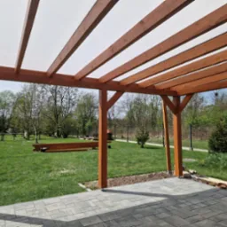 Drewniana pergola z transparentnym zadaszeniem, widok z poziomu tarasu z kostki brukowej na zielony ogród z bramką piłkarską w tle.