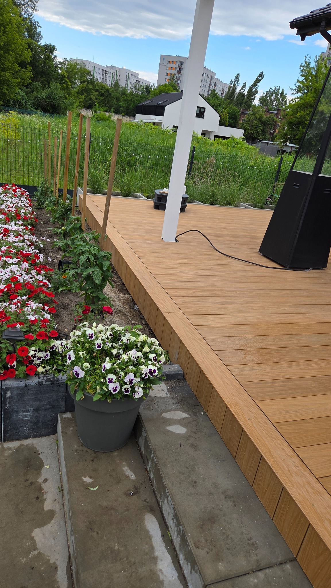 Jasny taras kompozytowy z donicami kwiatów, widok z bliska. Elementy ogrodowe, w tle zieleń i zabudowa miejska. Słup pergoli.