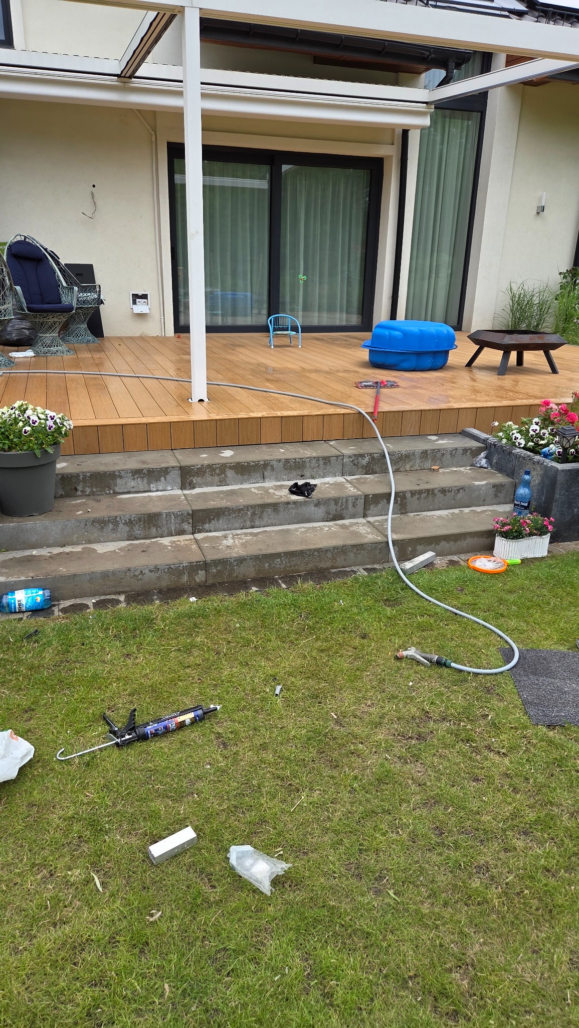 Montaż tarasu kompozytowego w trakcie: widoczne narzędzia, klej, deski tarasowe i węże do czyszczenia na trawie. Detal z bliska, zieleń w tle.