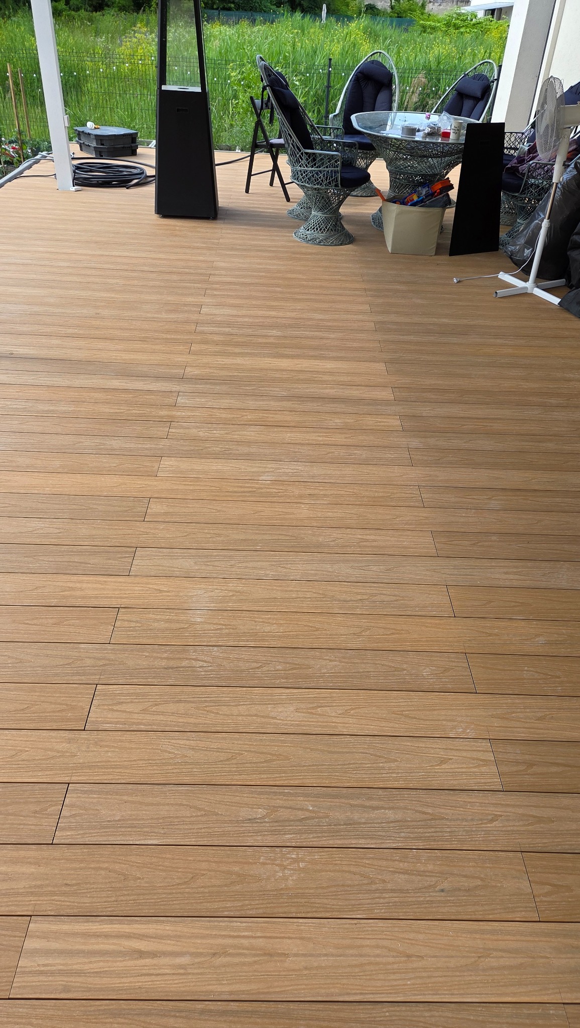 Beżowy taras z desek kompozytowych, z widocznymi detalami faktury drewna. W tle meble ogrodowe i zieleń, tworzące przytulną przestrzeń wypoczynkową.