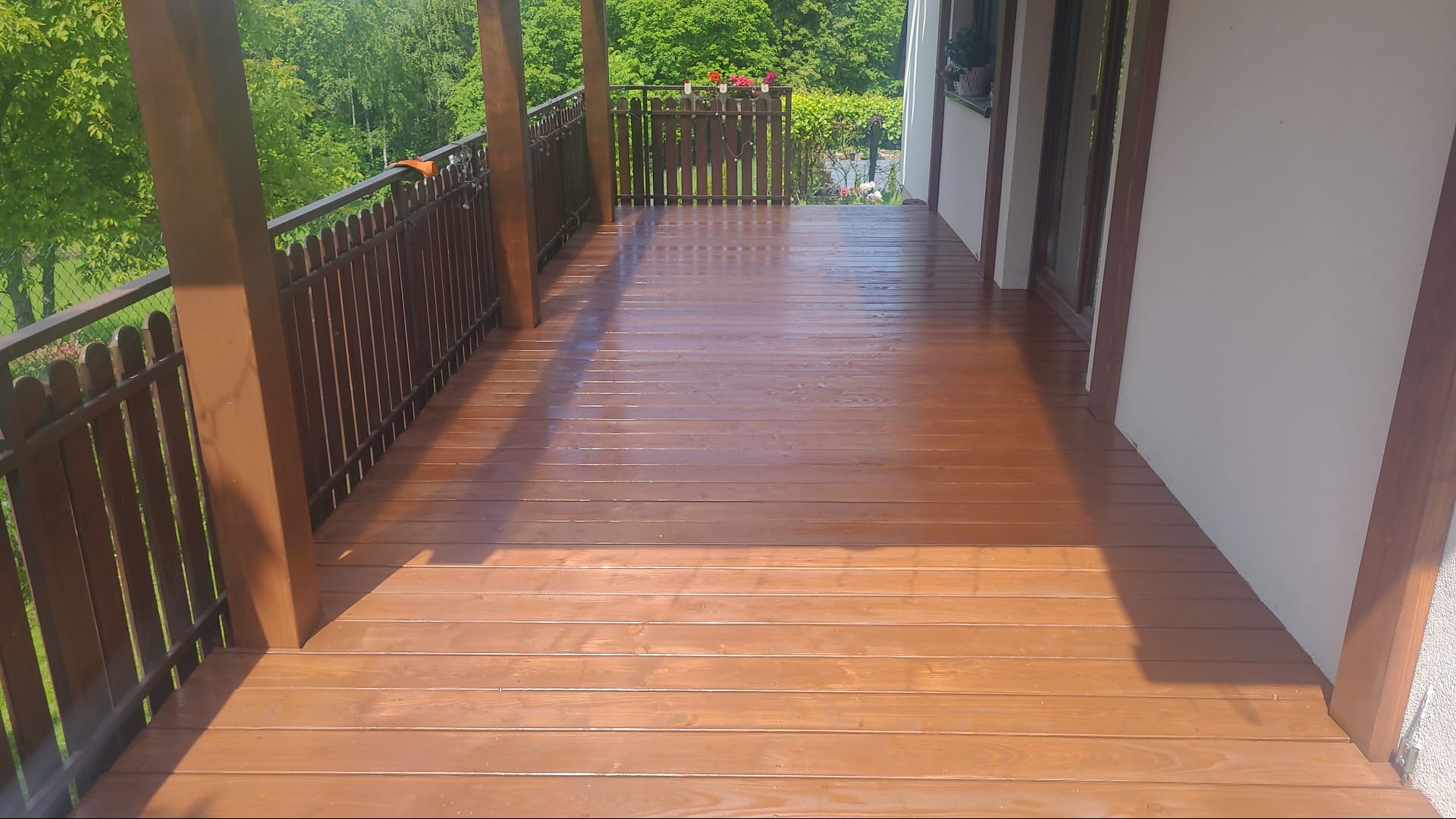 Błyszczący, brązowy taras z drewnianą balustradą, widok na zieleń. Świeżo polakierowana podłoga tarasu w promieniach słońca. Elementy drewniane konstrukcji.