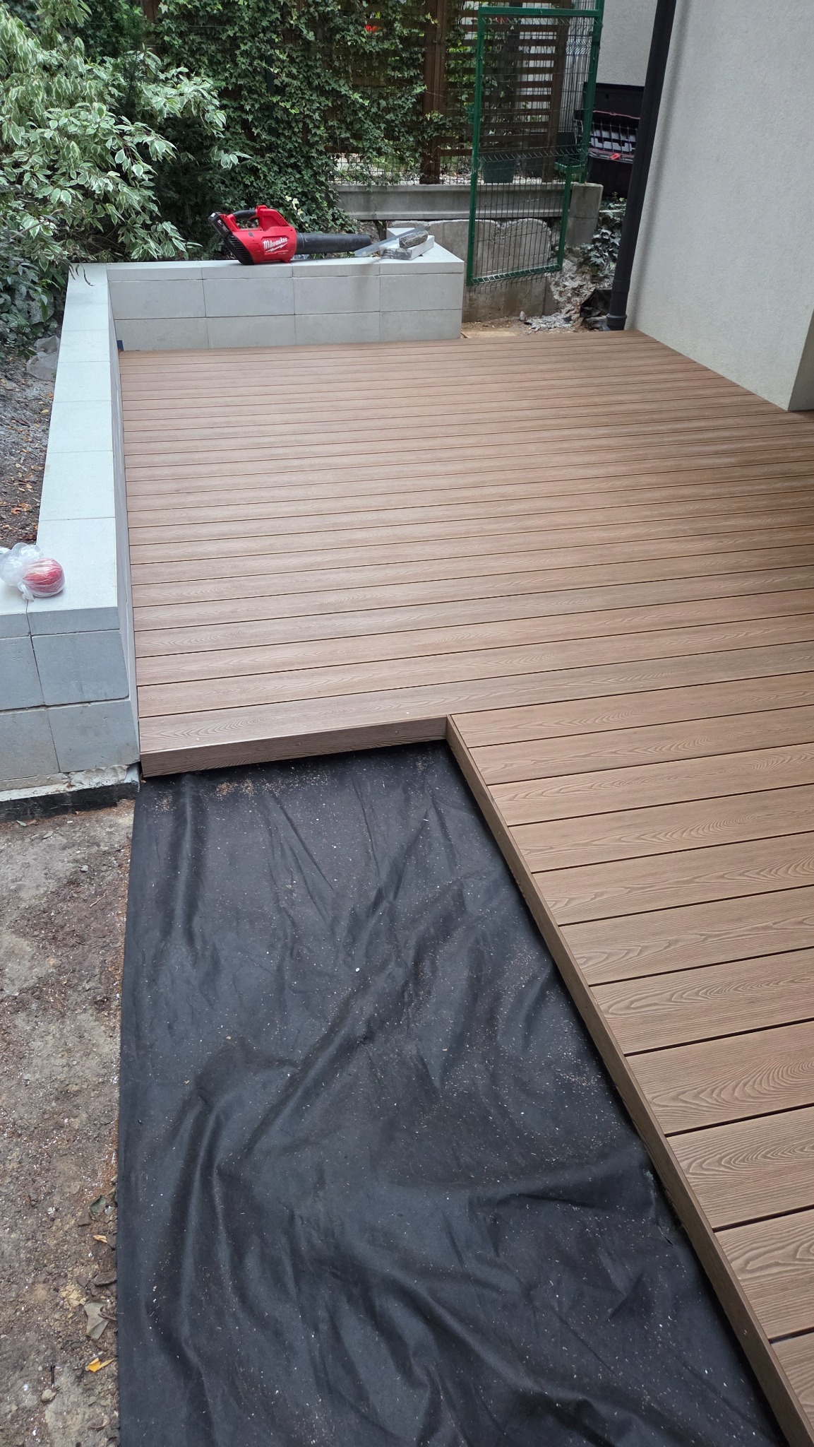 Montaż tarasu kompozytowego: widok z góry na deskę ułożoną na czarnej membranie, z narzędziem (dmuchawa) na murku w tle. Kątowe ułożenie desek.